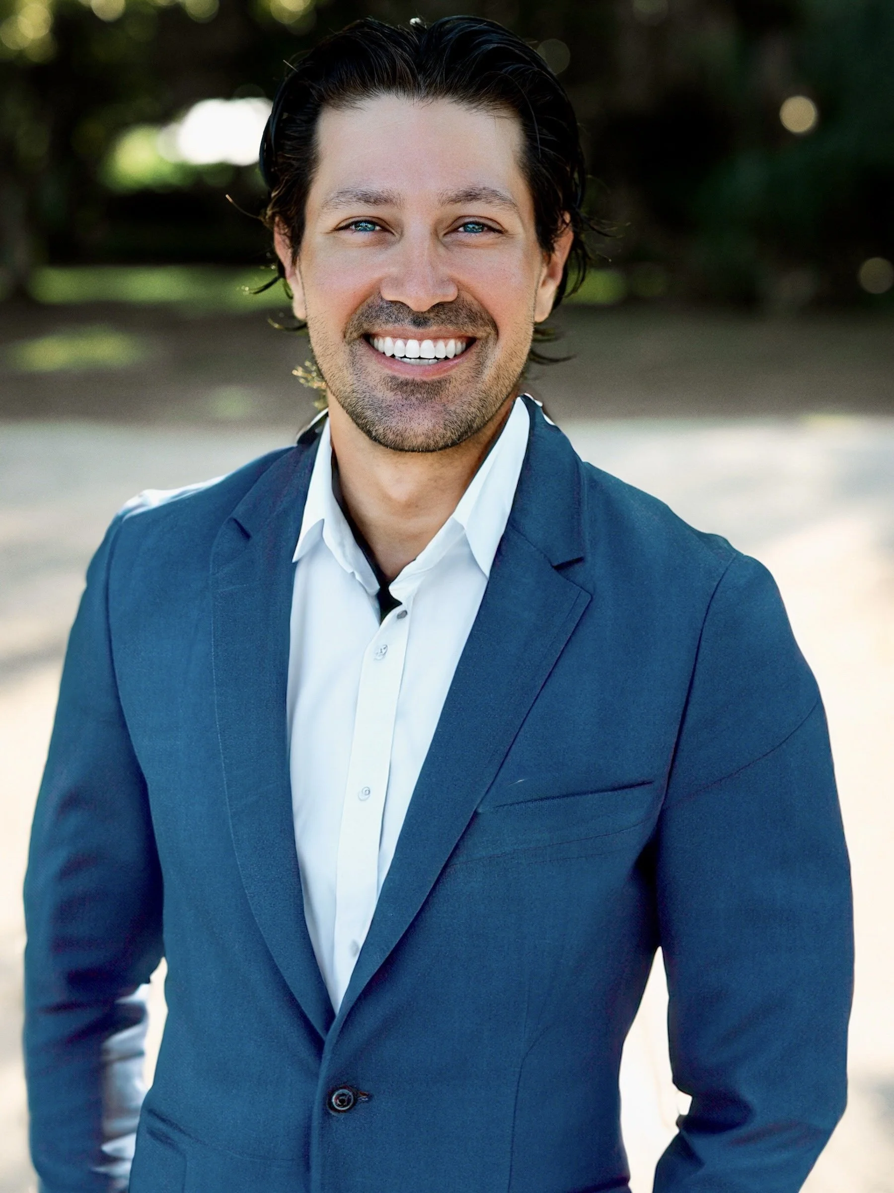 A man with short dark hair, bright blue eyes, and a beard, dressed in a dark blue suit with a white shirt, smiling outdoors with a blurred natural background.
