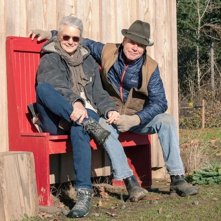 Mike Etzel and Carey Critchlow, owners of the Sequitur Vineyard in the Ribbon Ridge AVA, sitting together on a red wooden bench outdoors on a sunny day.
