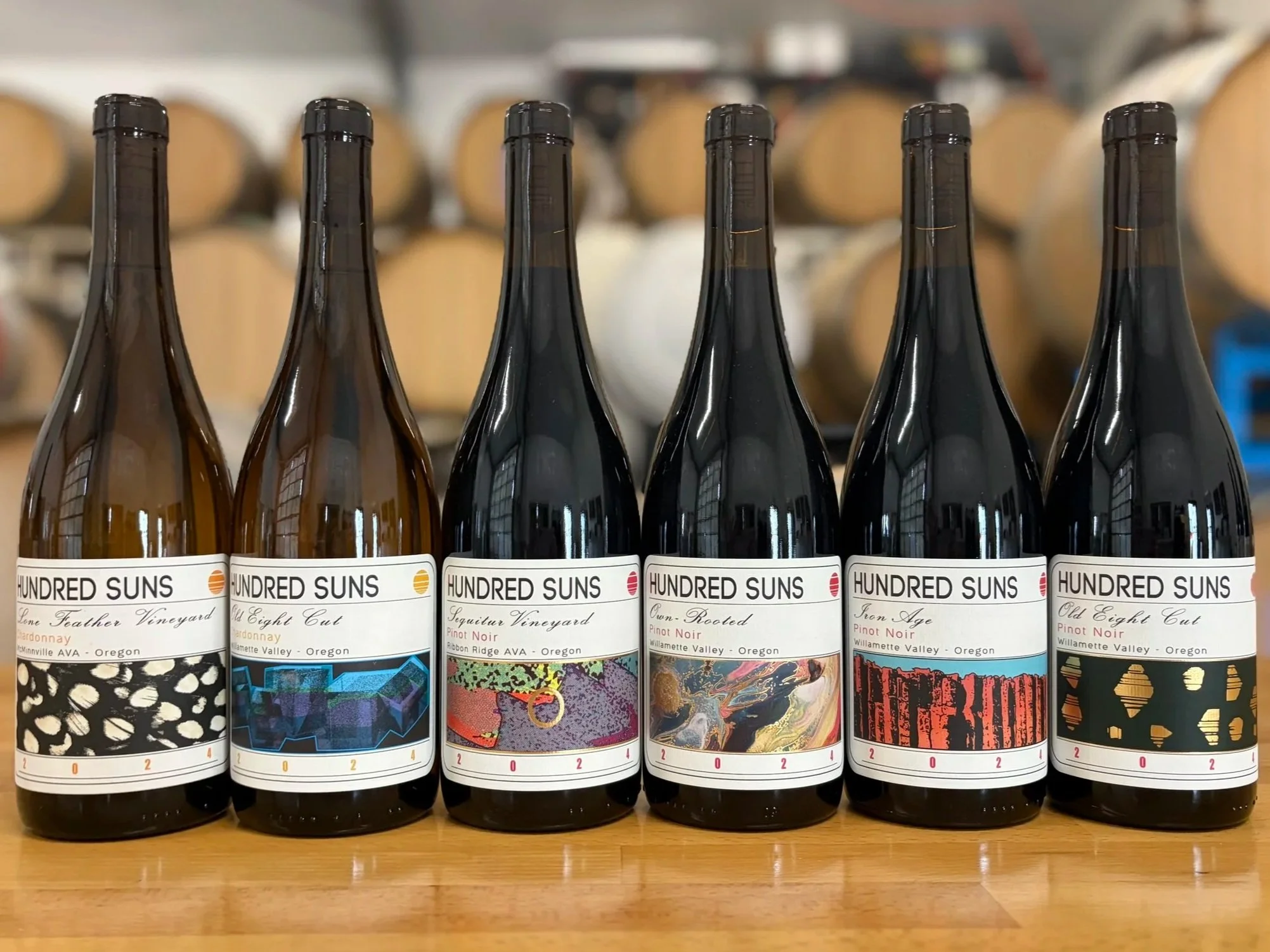 Row of wine bottles with labels from the winery Hundred Suns, featuring various artistic designs and names of different wine varieties, on a wooden surface.