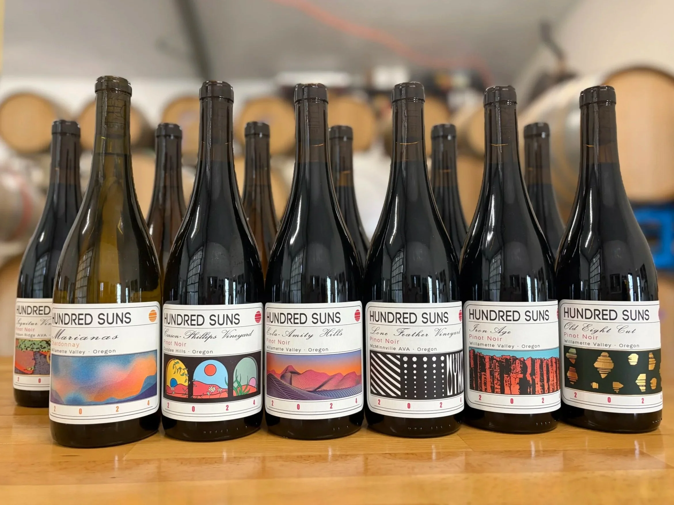 Multiple bottles of wine with colorful labels arranged on a wooden surface, with wooden wine barrels in the background.