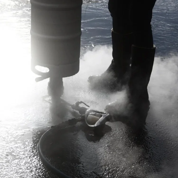 Person in black boots using a propane torch to melt or cut asphalt on pavement, with steaming or smoky dispersion around.