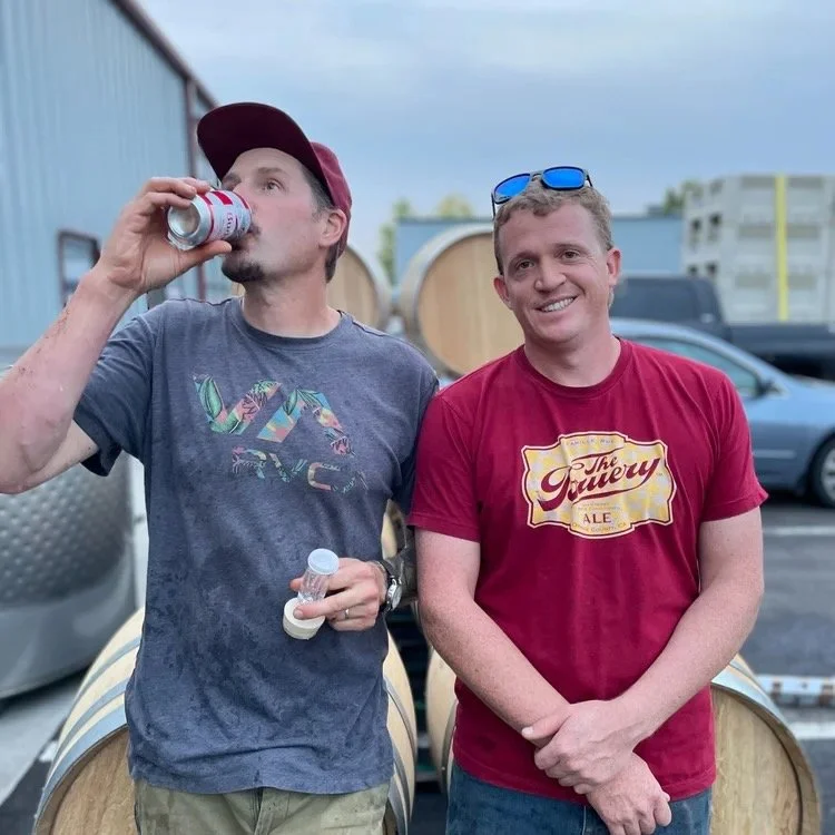 Grant Coulter and Grant Phillips of Carson Phillips Vineyards in the Dundee Hills AVA standing outdoors near wine barrels; one drinking from a can, the other smiling with sunglasses on his head.