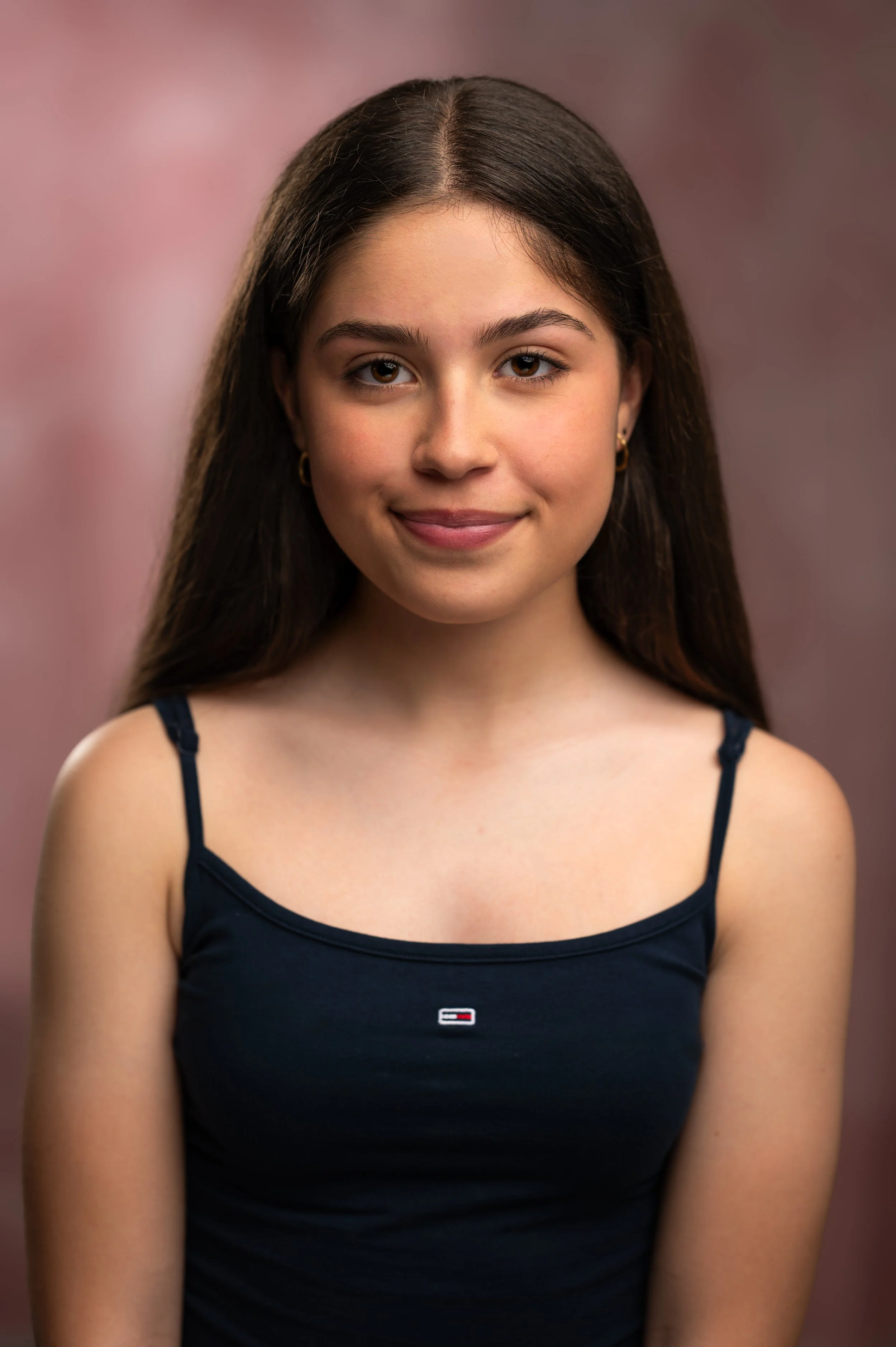 Close-up portrait of a young woman with long brown hair, wearing a navy blue tank top, smiling softly against a pink blurred background.