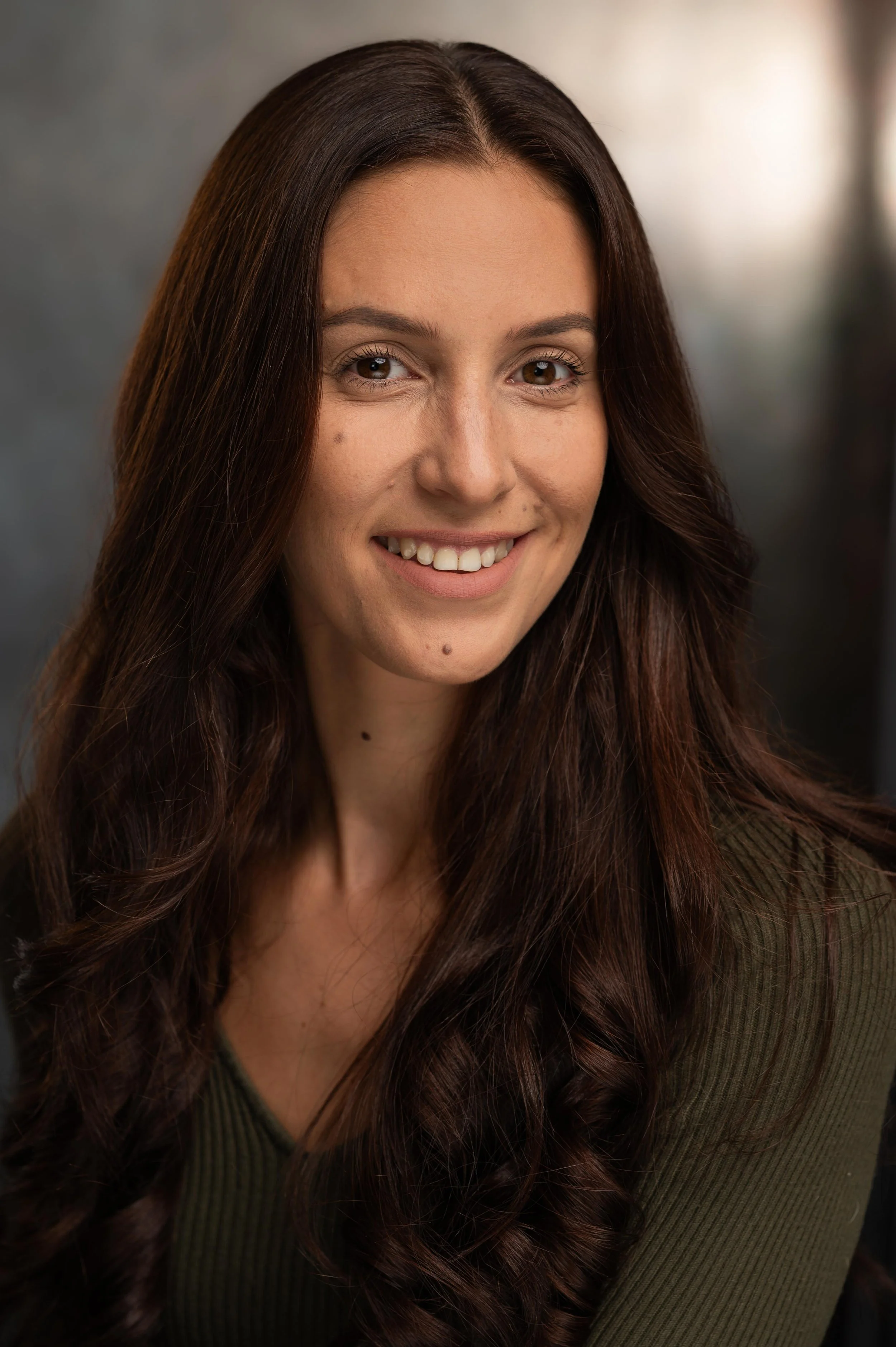 Close-up of a smiling woman with long, wavy brown hair, wearing a dark green top, against a blurred background.