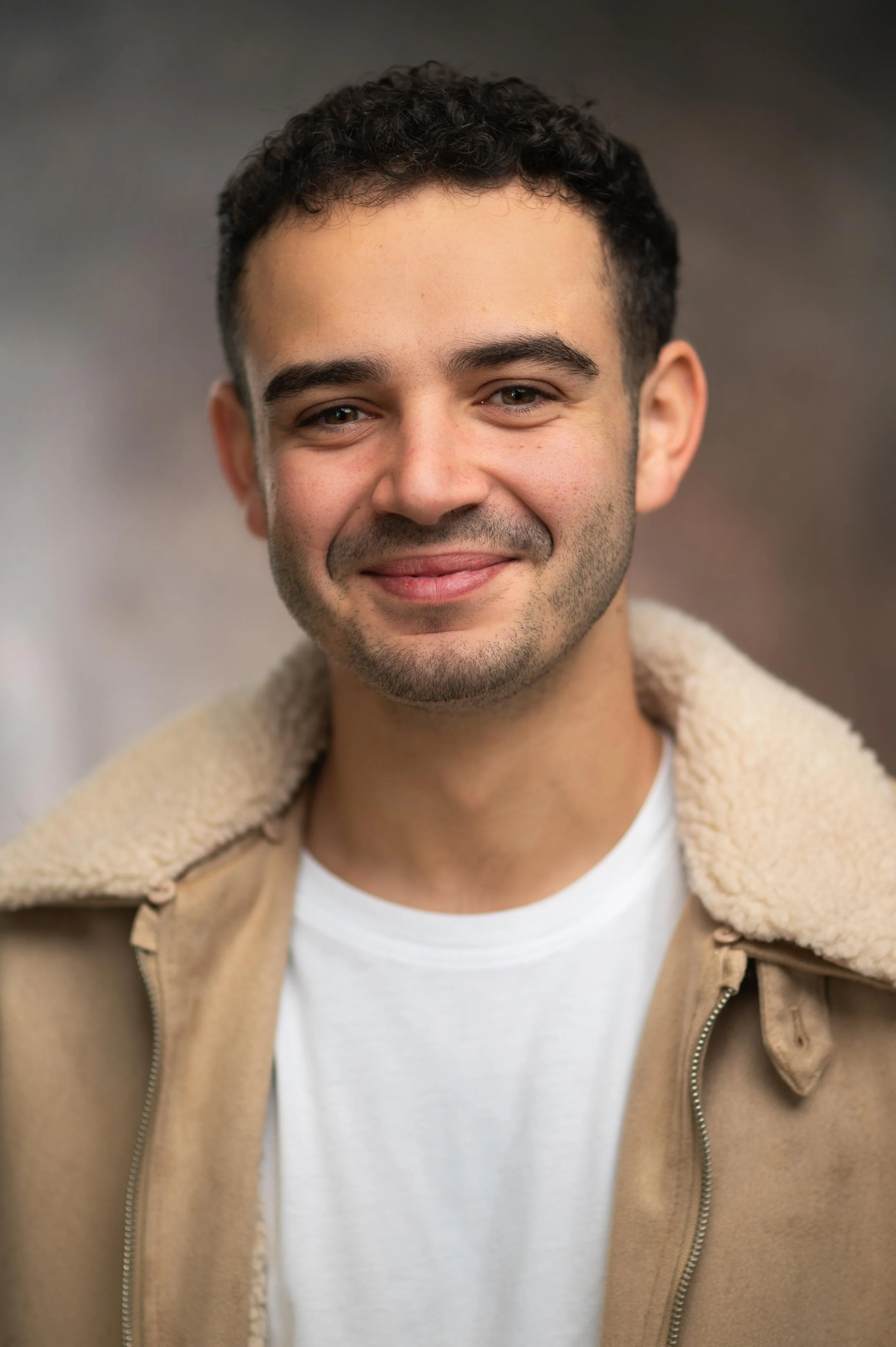 Close-up photo of a smiling Jamie Cardona man with short, curly dark hair wearing a beige jacket with a shearling collar over a white t-shirt, against a neutral blurred background.