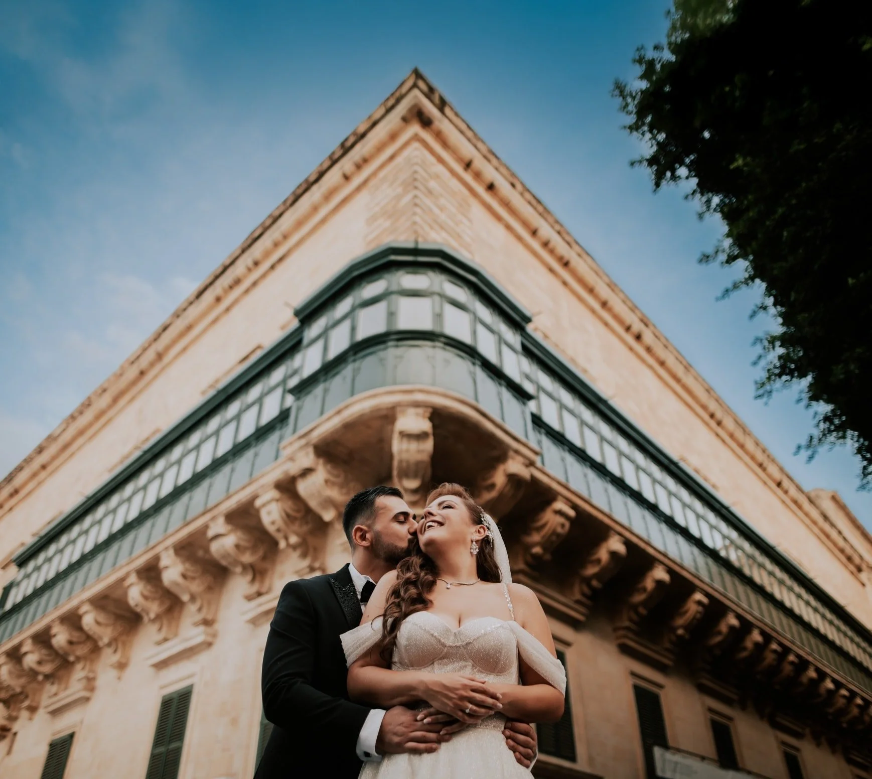 A bride and groom stand close together under an ornate historic building, sharing a kiss and looking happy on their wedding day.