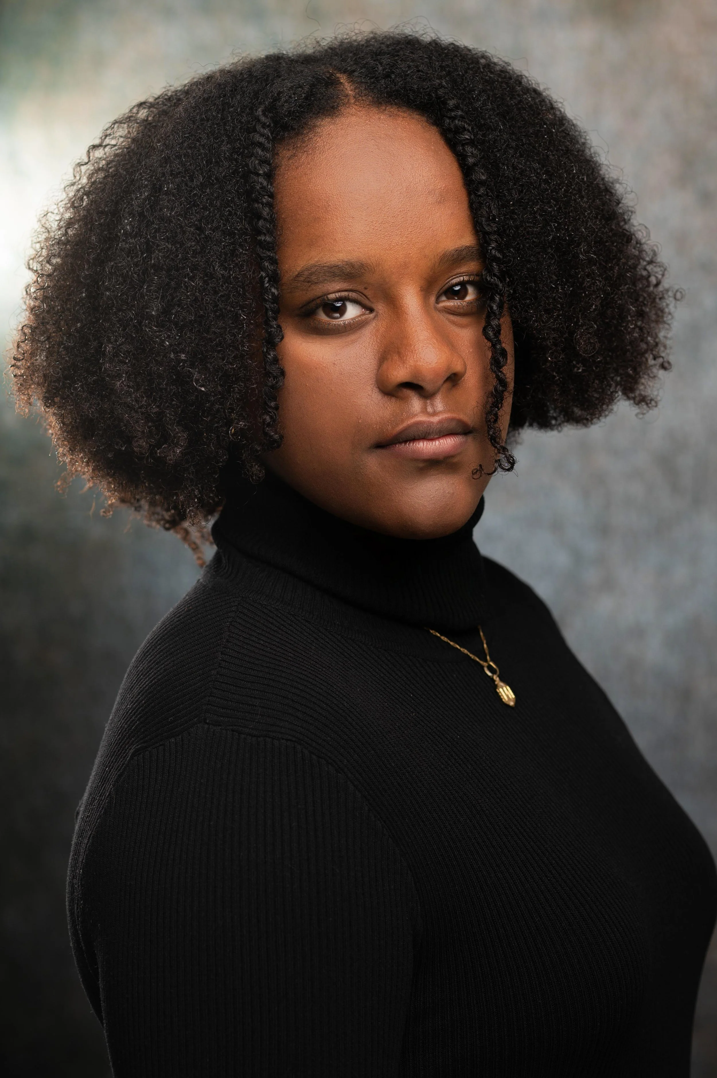 A portrait of a young woman with curly hair wearing a black turtleneck and a gold necklace, looking at the camera with a neutral expression.