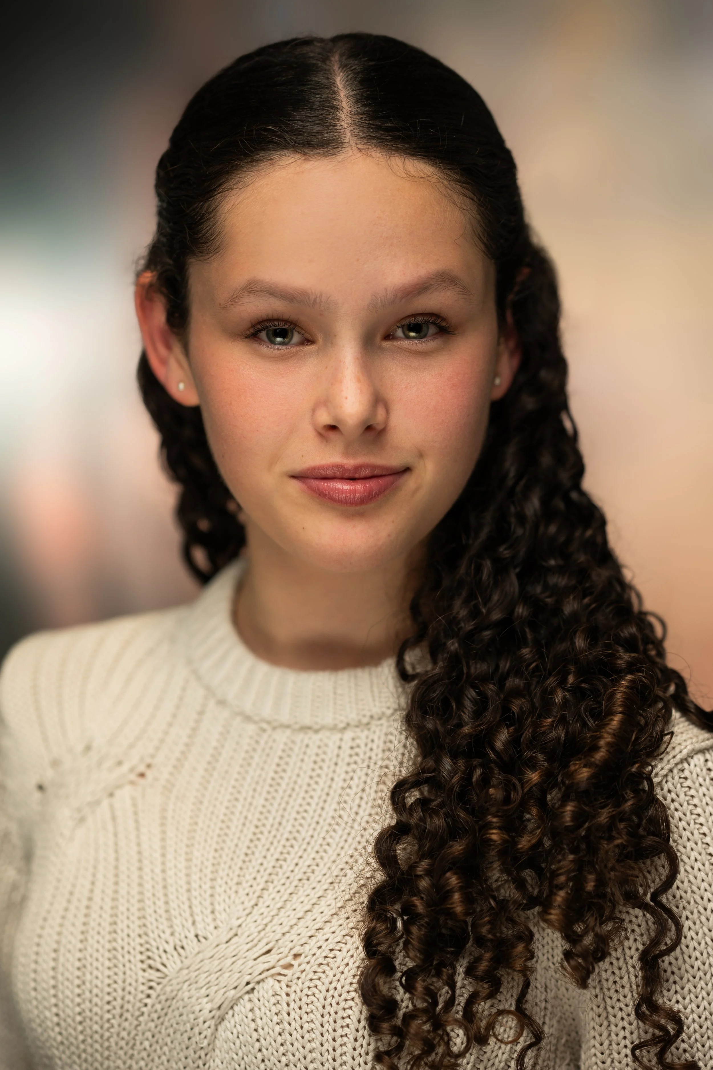 A young woman with long, dark curly hair, wearing a cream-colored sweater, looking at the camera with a slight smile.