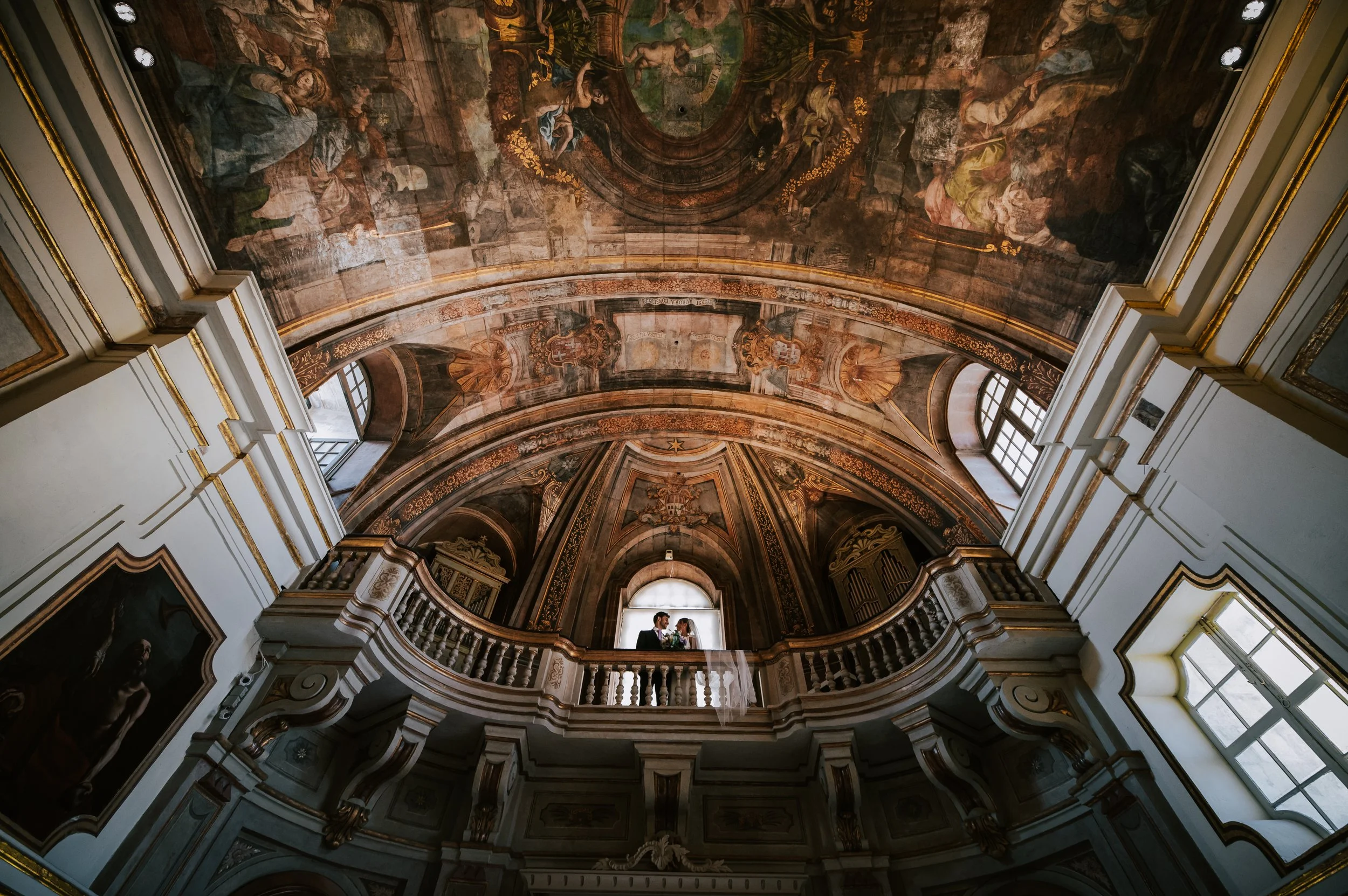 A couple in wedding attire stands on a balcony inside a grand, ornately decorated church with a high ceiling featuring intricate frescoes and gold trim.