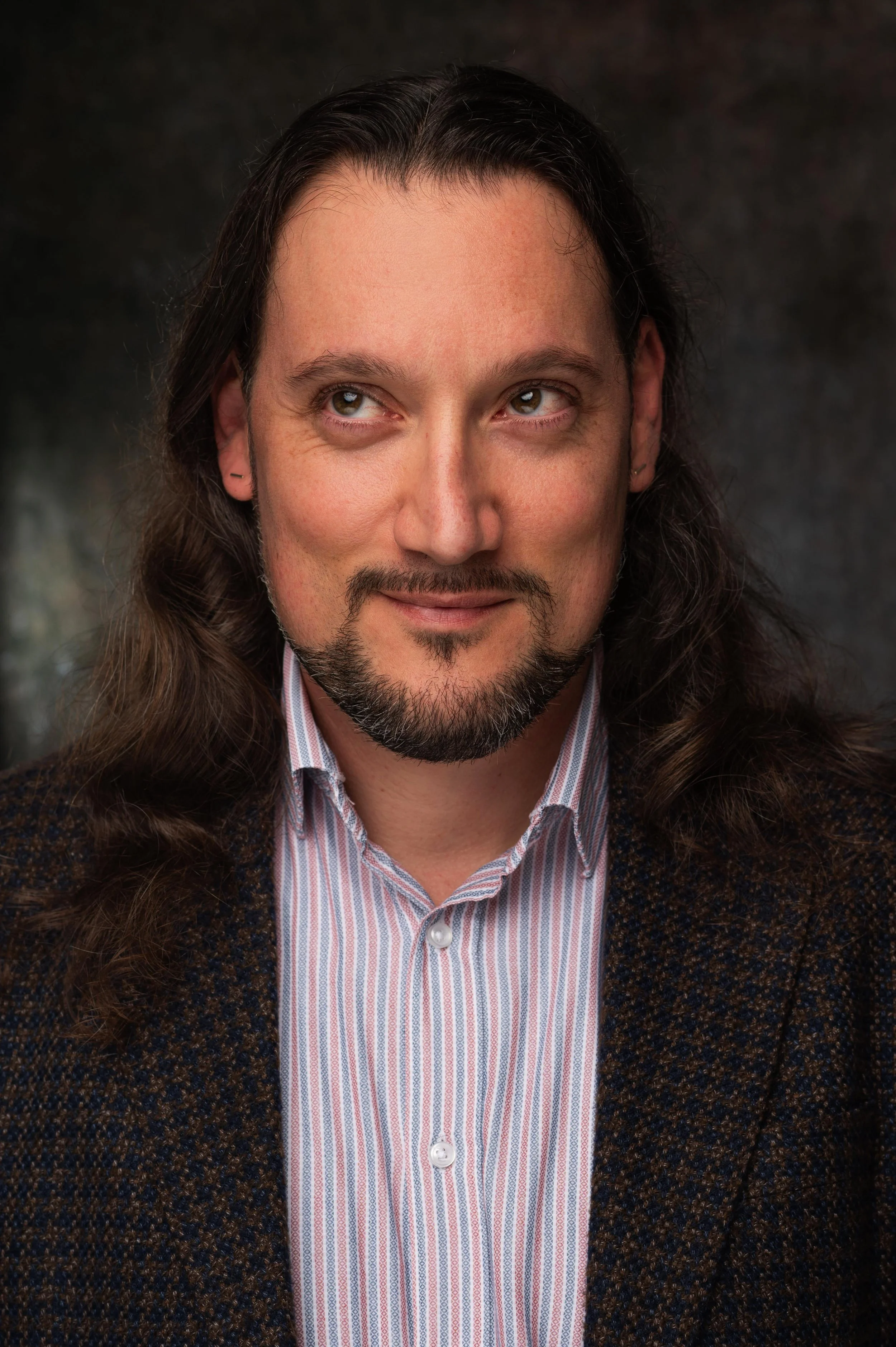 A man with long brown hair and a goatee, wearing a striped dress shirt and a dark, patterned blazer, looking off to the side with a slight smile.