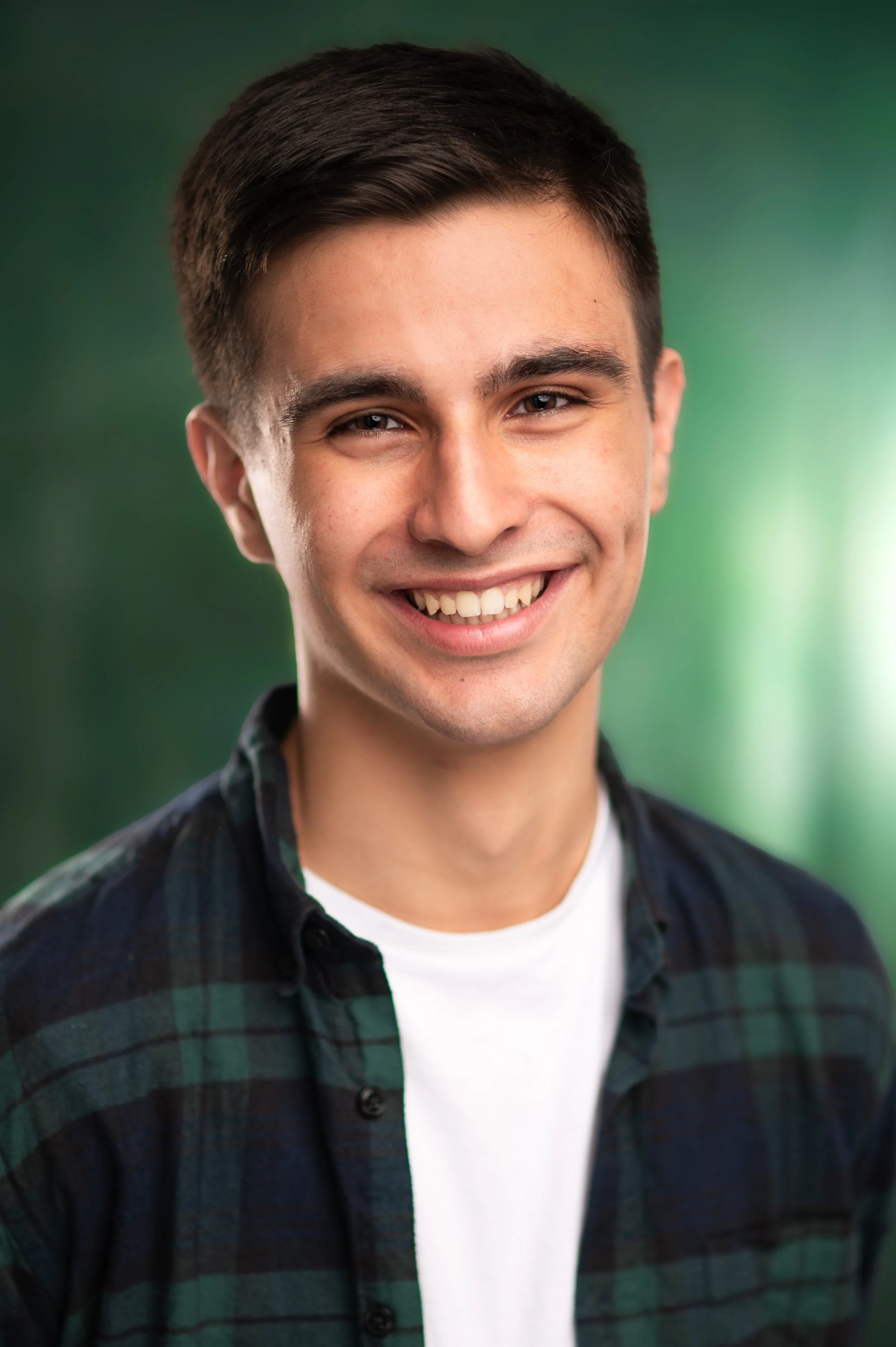 A young man with dark hair and a smile, wearing a white t-shirt and a dark green and navy plaid shirt, standing outdoors against a blurred green background.
