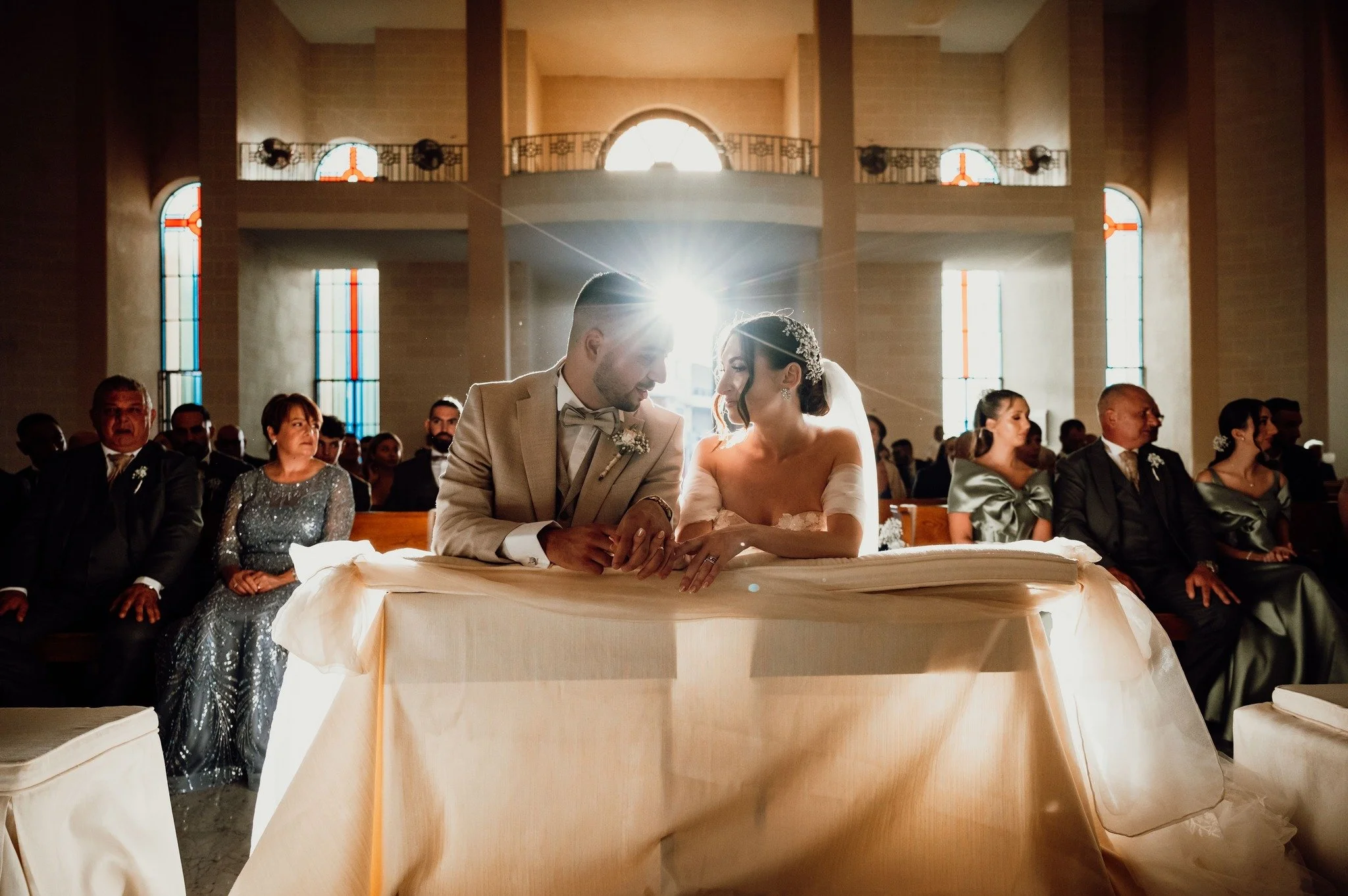 A bride and groom sitting at the altar during their wedding ceremony inside a church with stained glass windows, surrounded by seated guests.