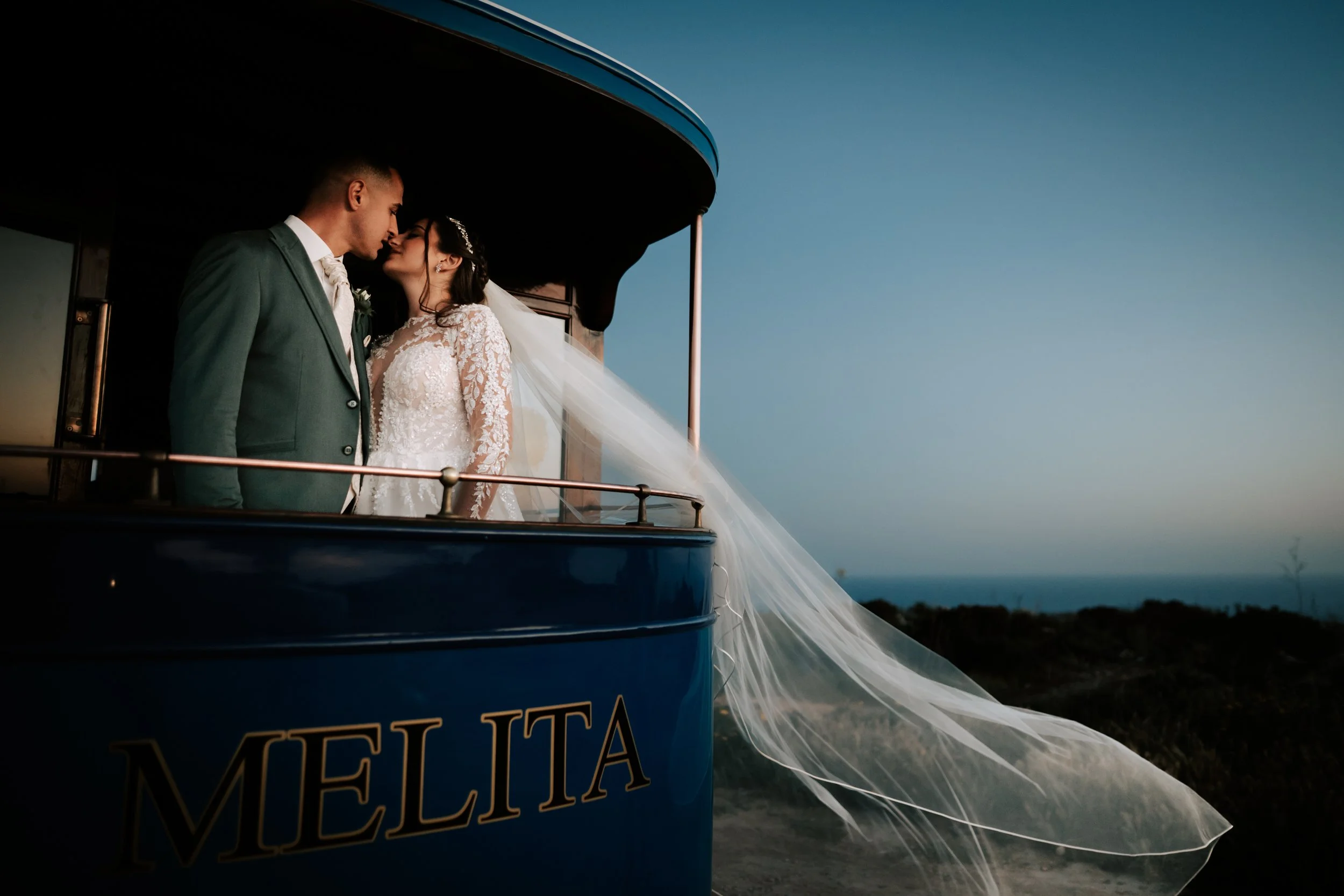 A bride and groom sharing a kiss inside a blue carriage with the word MELLIA on it, during sunset or dusk against a clear sky.