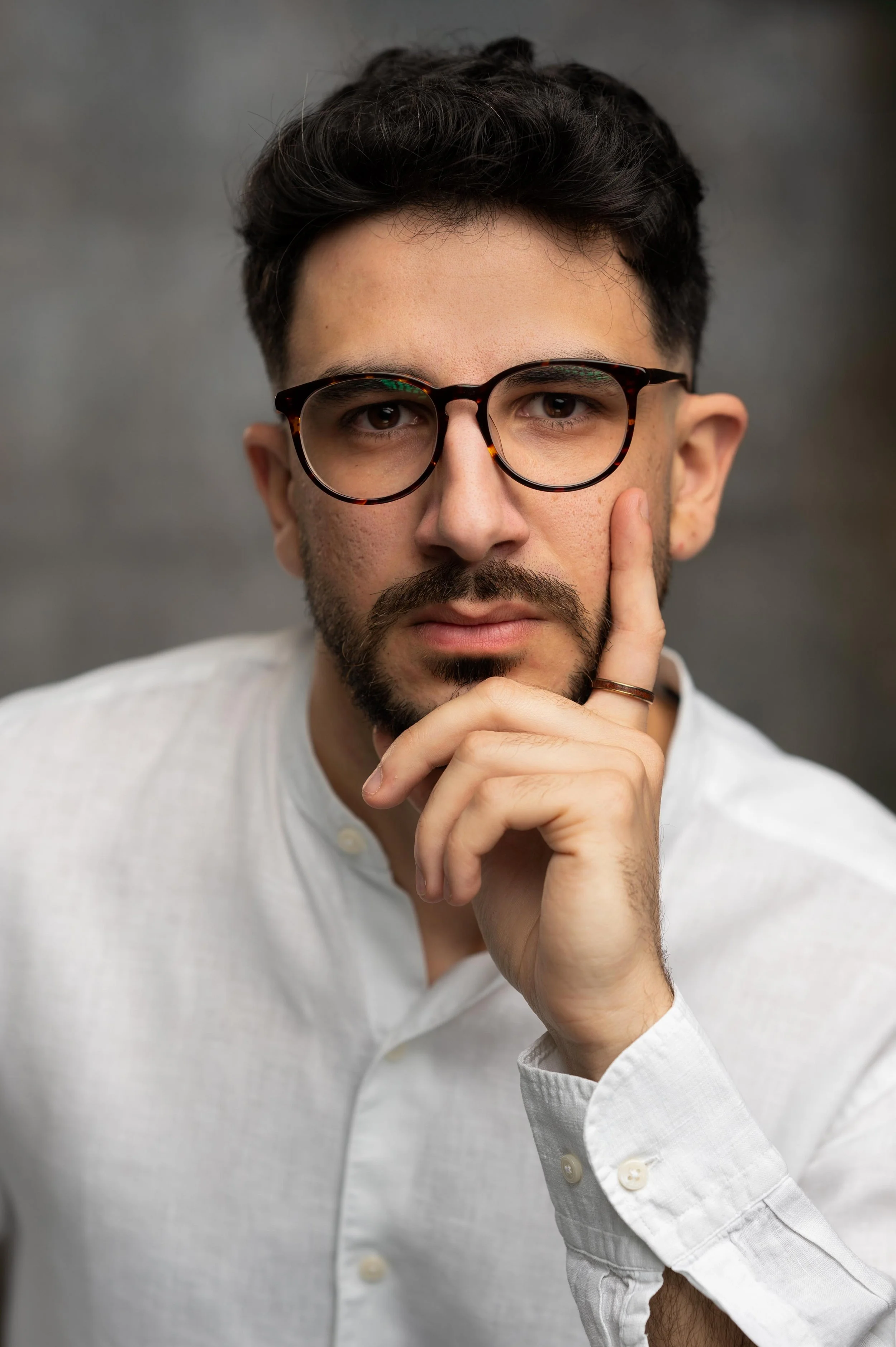 A man with dark hair, glasses, and a beard, resting his chin on his hand, wearing a white shirt through a thoughtful pose against a neutral background.