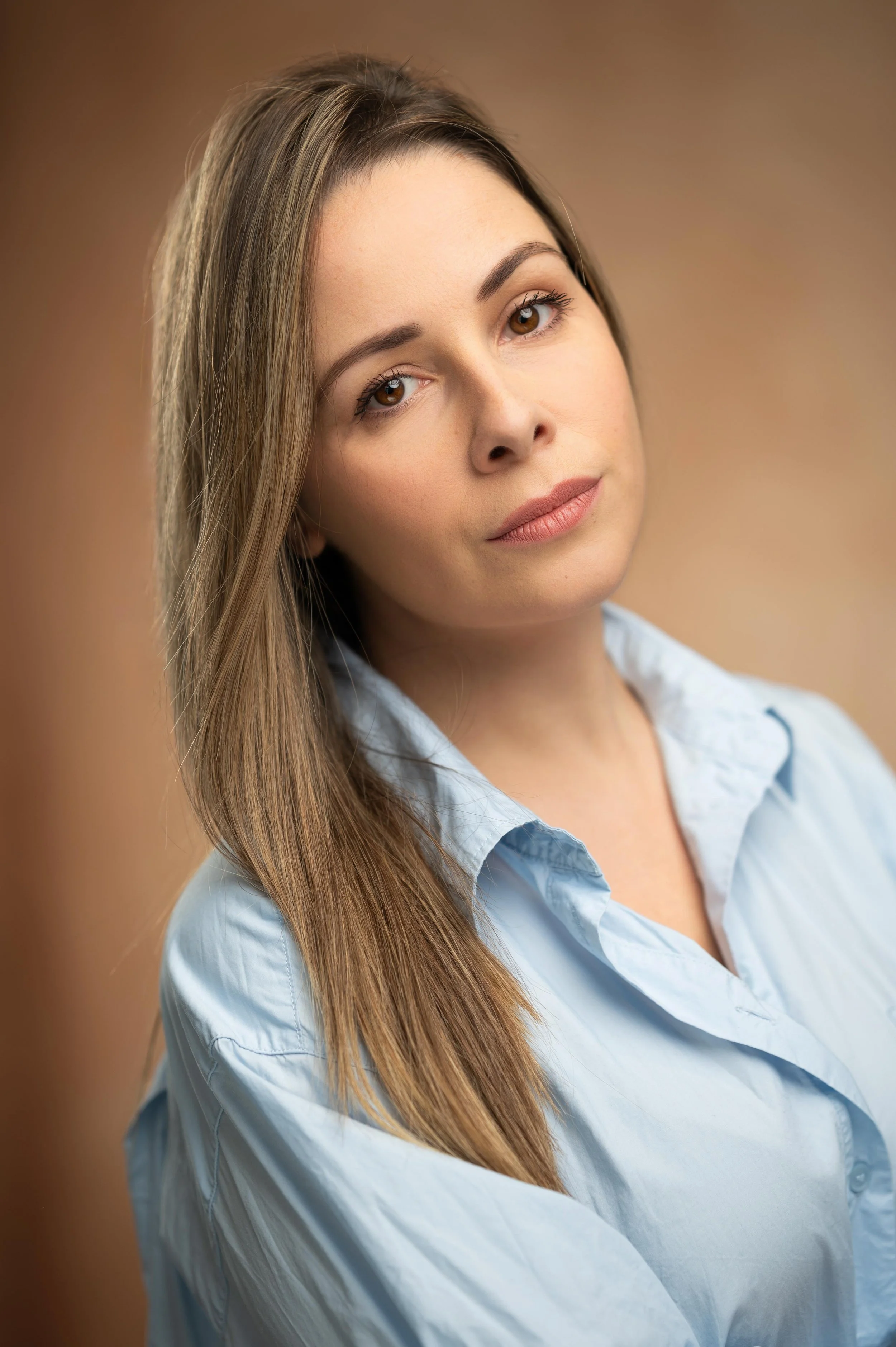 Close-up photo of a young woman with long brown hair, fair skin, wearing a light blue button-up shirt, looking softly at the camera against a blurred warm brown background.
