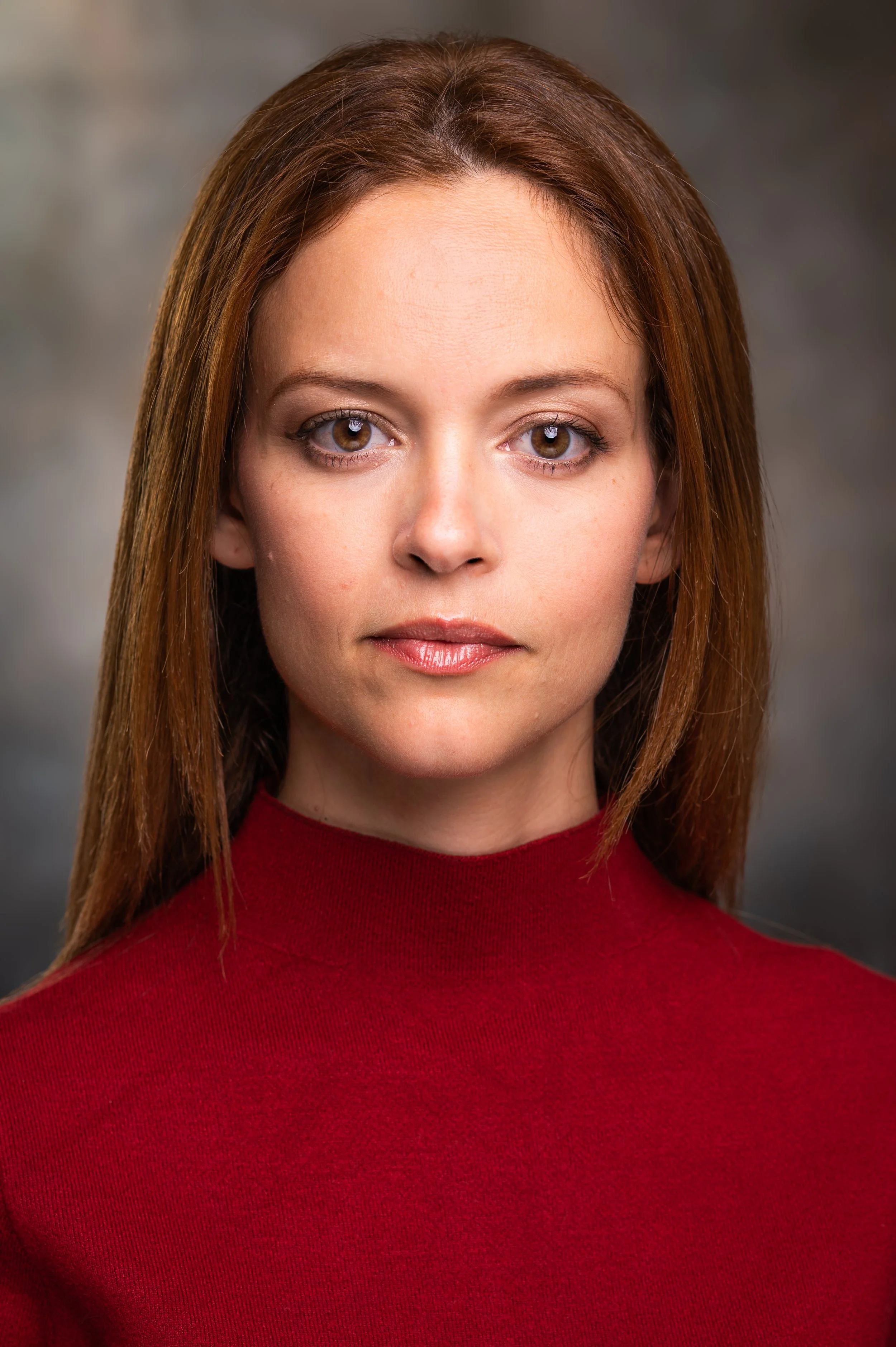 Close-up of a woman with brown hair, wearing a red top, with a neutral expression, against a blurred gray background.