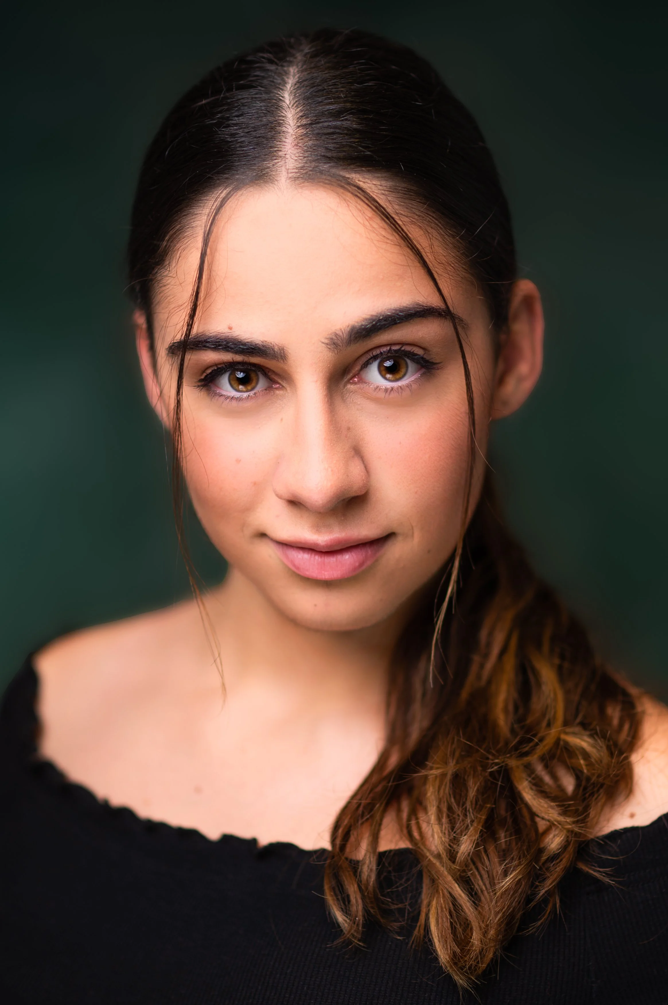 Close-up portrait of a woman with hazel eyes and dark brown hair with lighter brown curls, wearing a black top, against a dark green background.