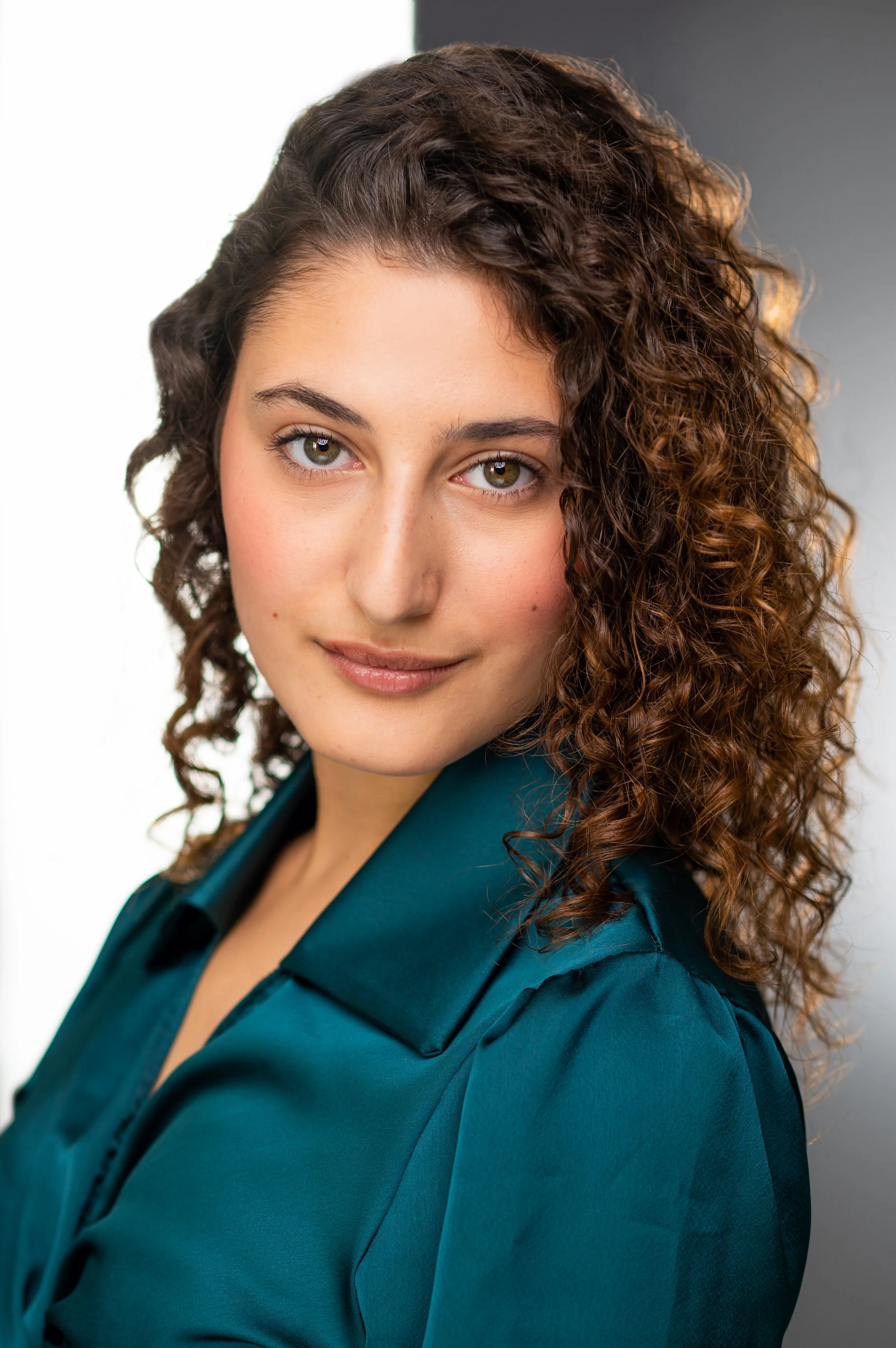 A young woman with curly brown hair and hazel eyes wearing a teal blazer, smiling slightly, against a neutral background.