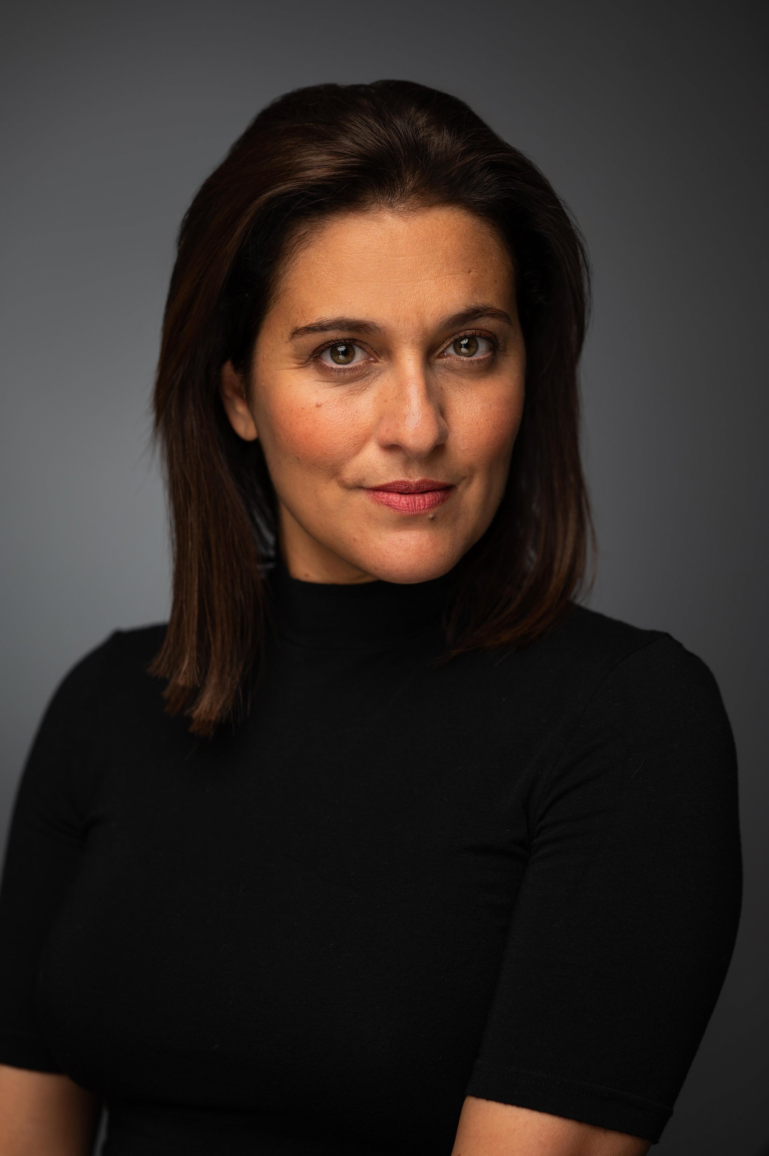 Close-up portrait of a woman with brown hair, wearing a black turtleneck, looking directly at the camera against a dark grey background.