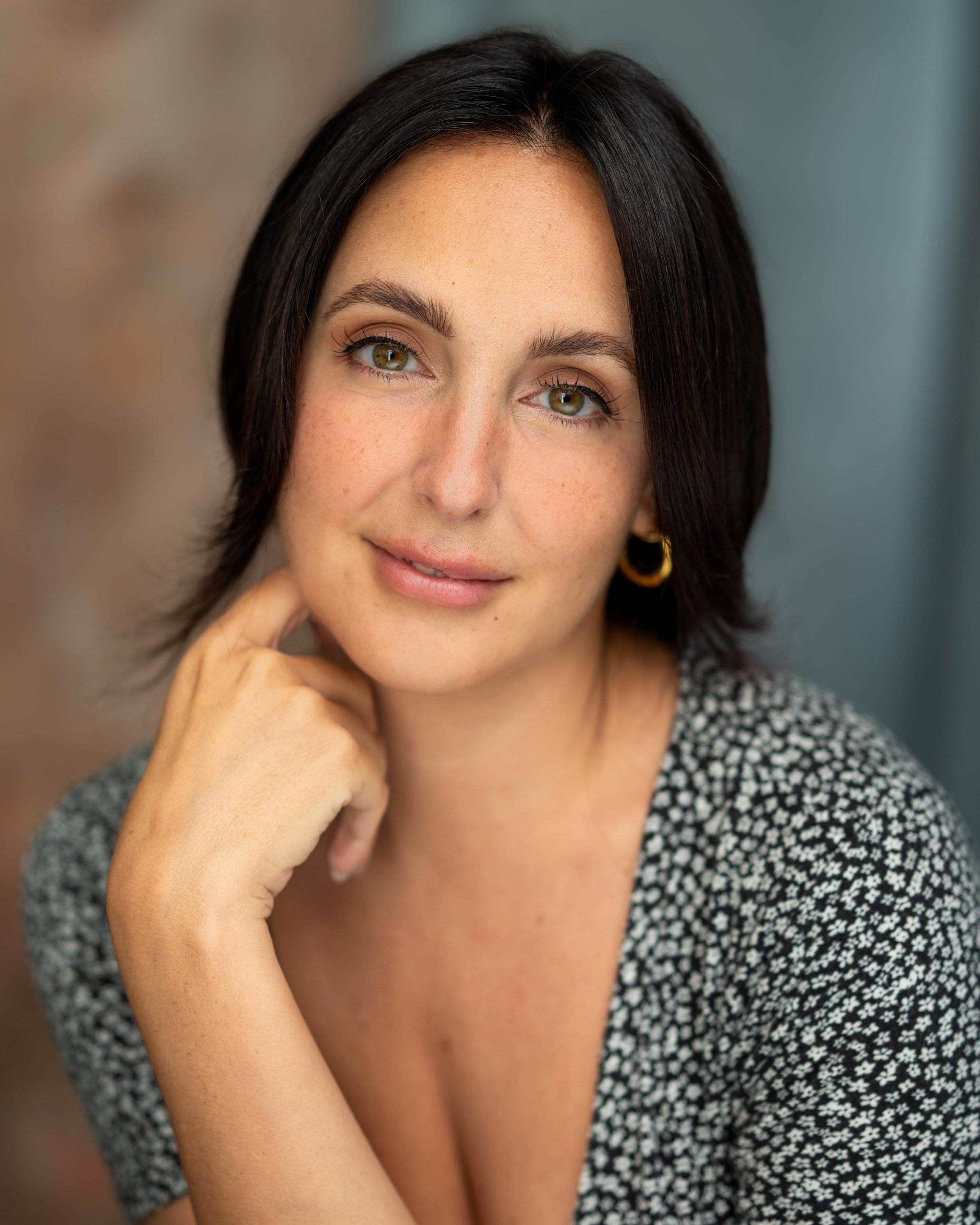 A woman with dark hair, green eyes, and wearing a black and white patterned top, smiling gently, resting her hand on her chin.