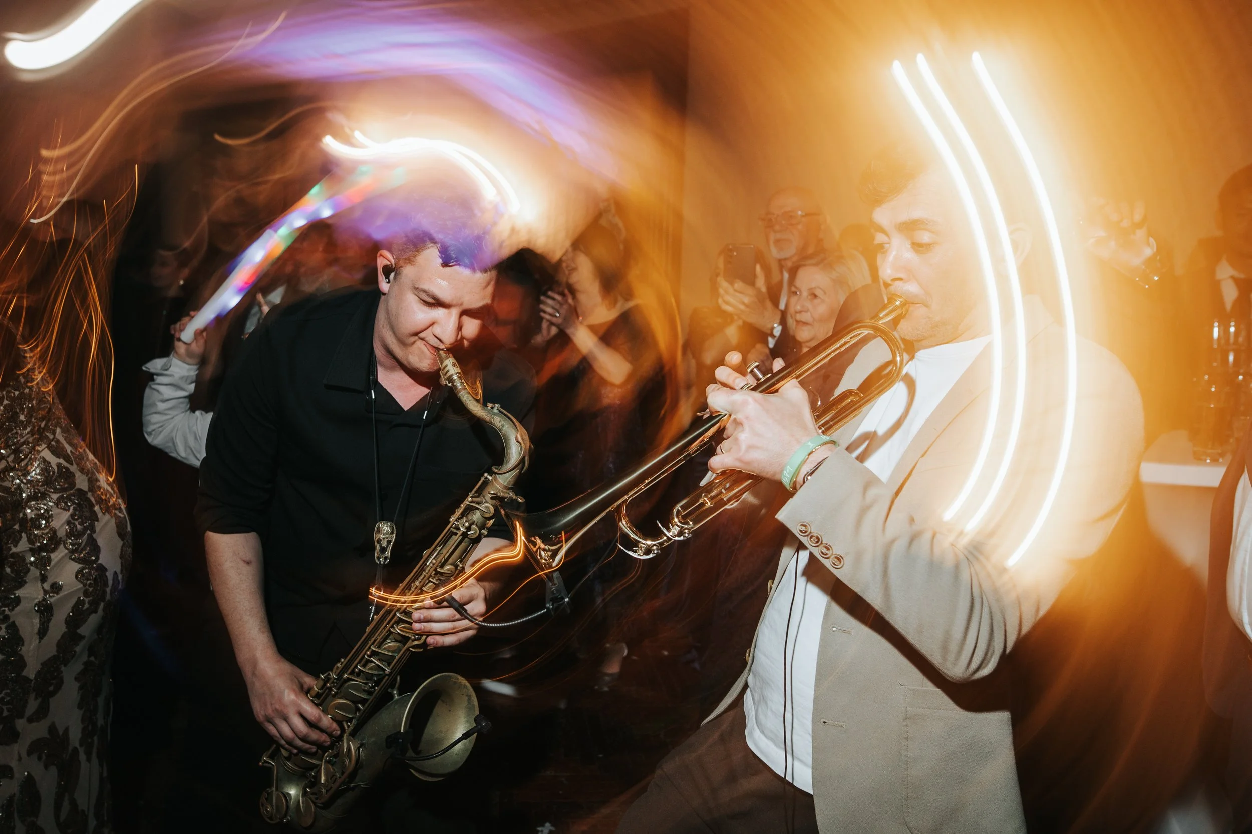 Two musicians playing saxophone and trumpet at a lively indoor event with colorful lights and an audience.