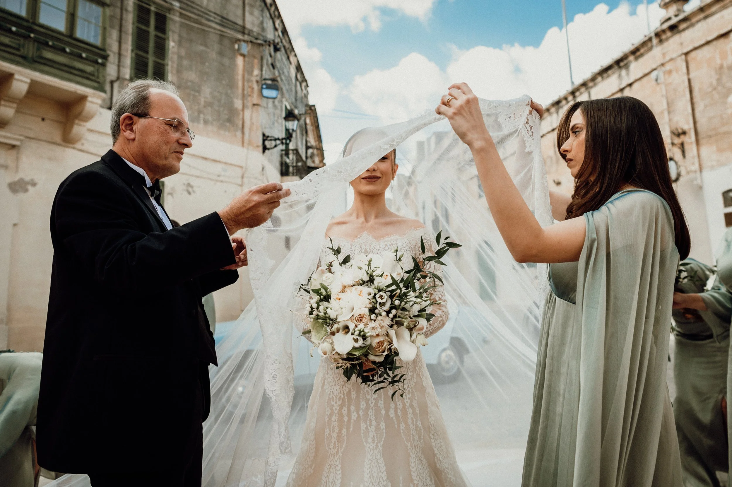A bride in a lace wedding dress holds a bouquet of white and blush flowers and is standing under a lace veil held by two women, with an older man in a tuxedo on her left and a woman in a light dress on her right. They are outside in front of old buil