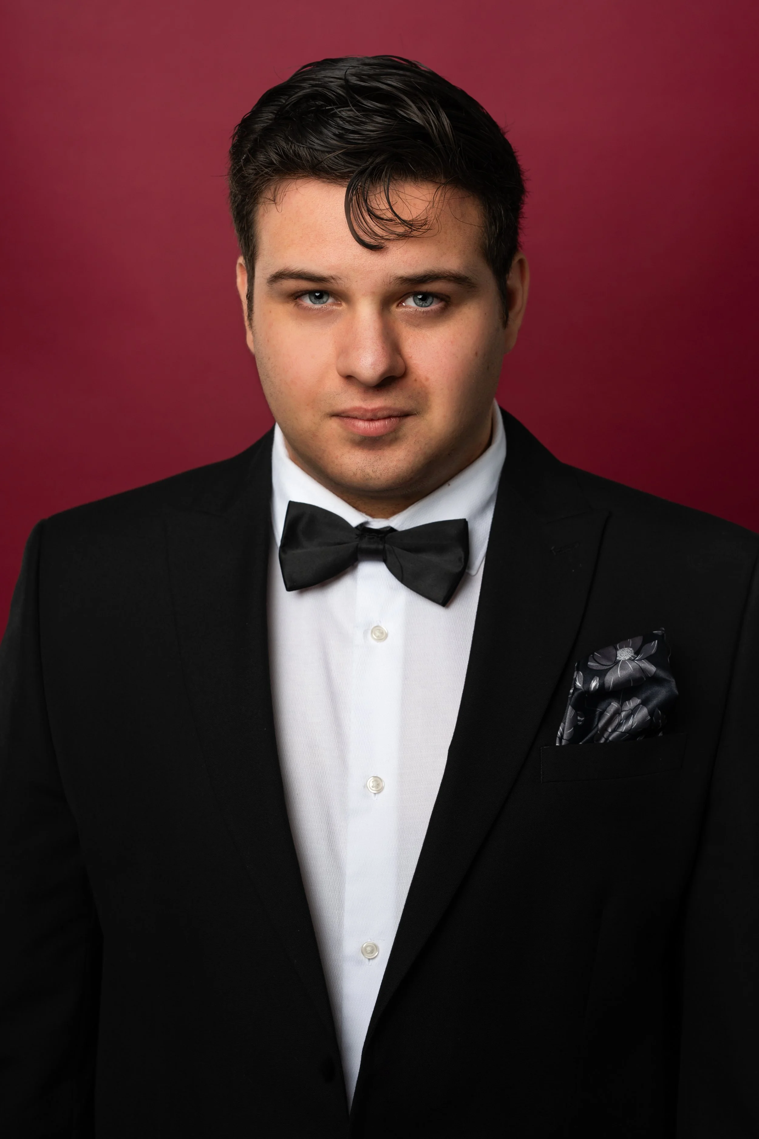 A young man dressed in a black tuxedo with a bow tie and a floral pocket square, against a red background.