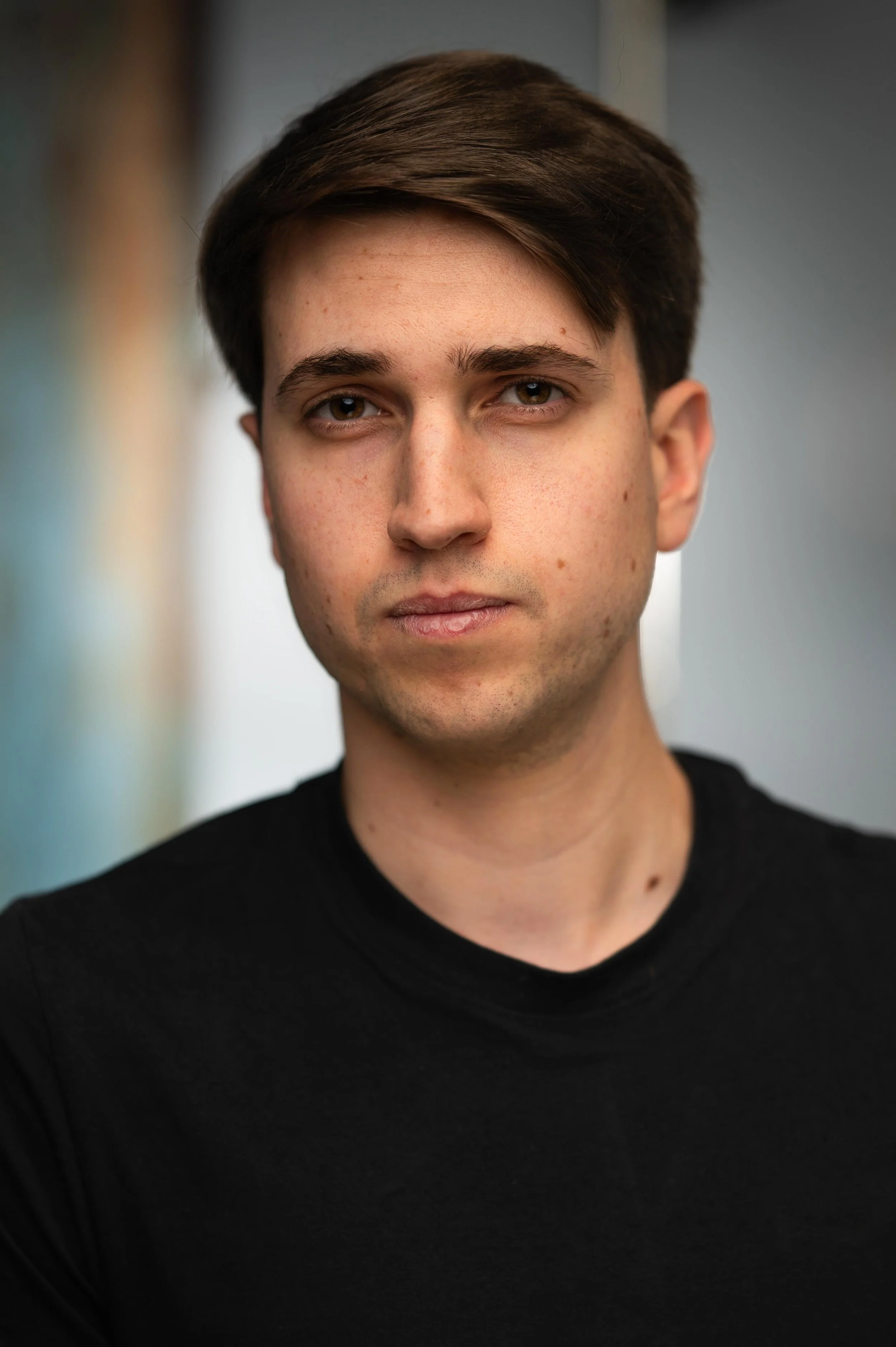Close-up of a young man with brown hair, wearing a black shirt, looking directly at the camera with a neutral expression. The background is blurred.
