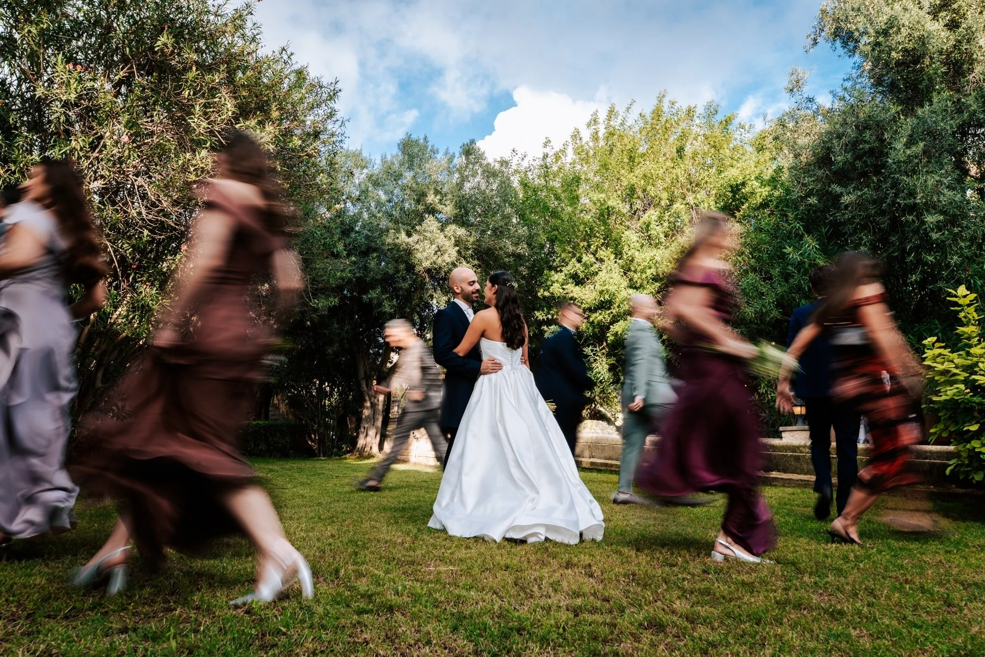 A bride and groom stand together smiling in a garden, surrounded by blurred guests walking past during a wedding celebration, under a blue sky with clouds.