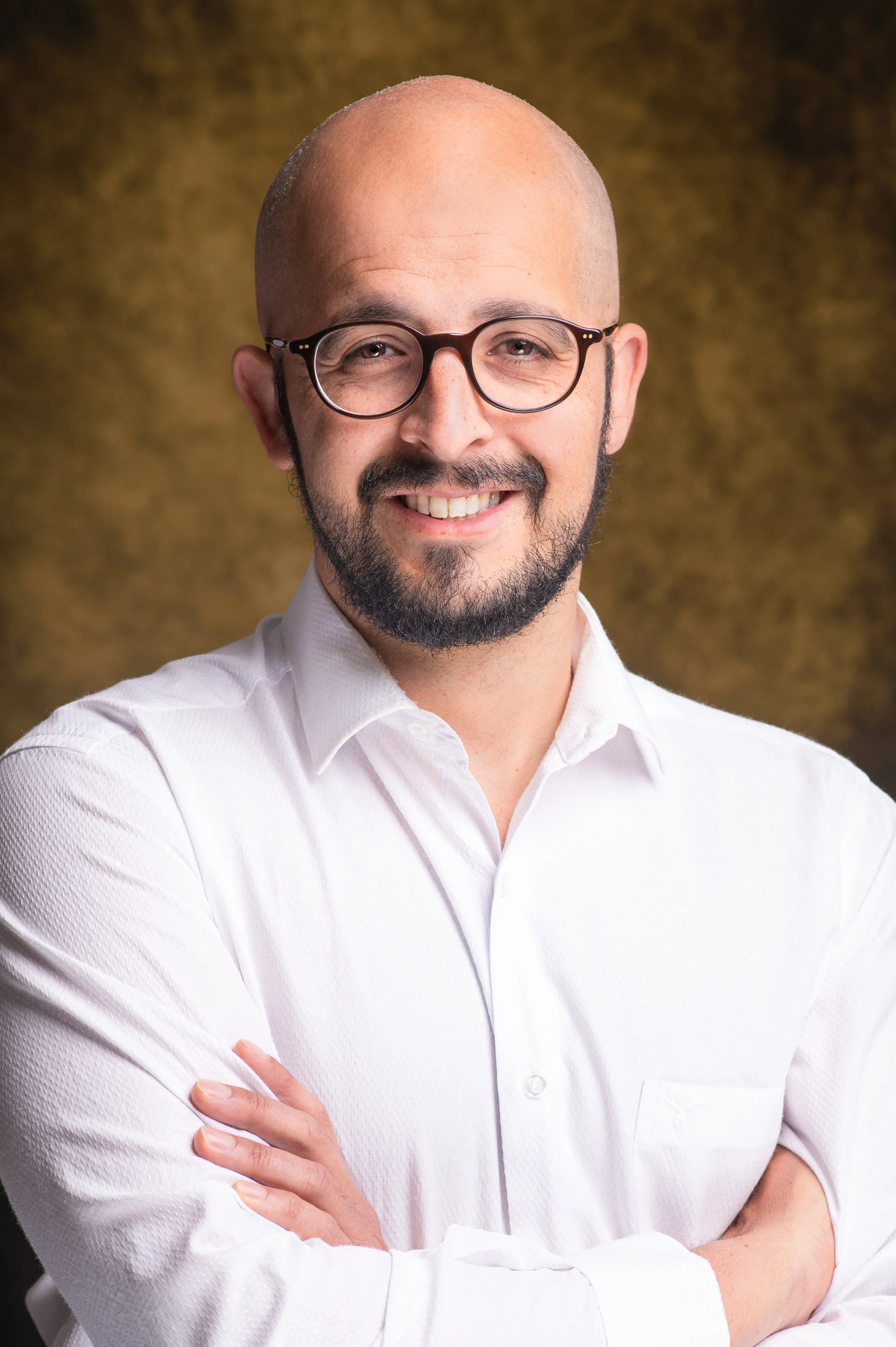 Bald man with glasses, beard, and mustache smiling, wearing a white dress shirt, with arms crossed, in front of a brownish background.