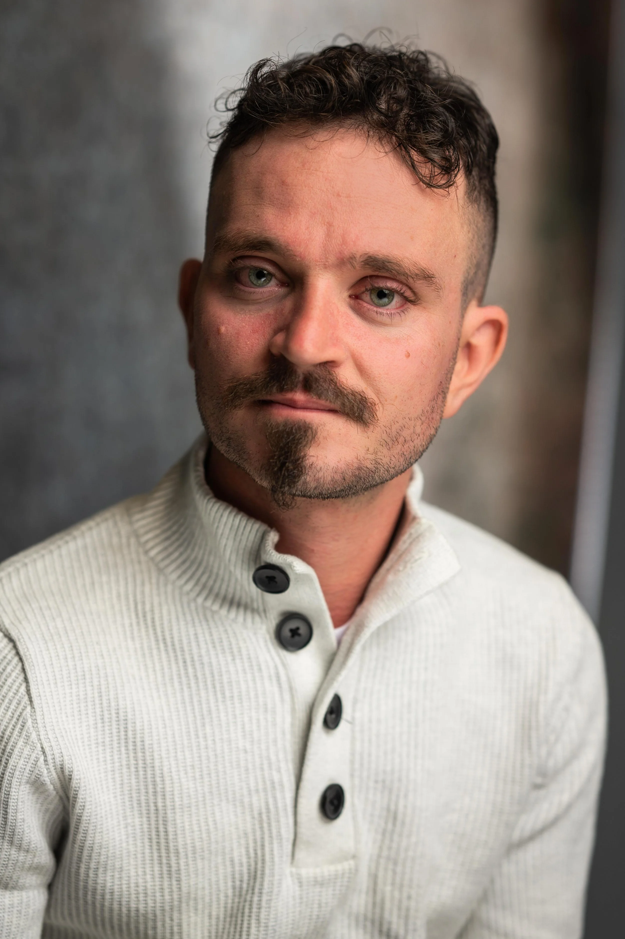 Close-up portrait of a man with light skin, short curly hair, and a mustache and goatee, wearing a cream-colored button-up sweater with black buttons, standing against a neutral background.