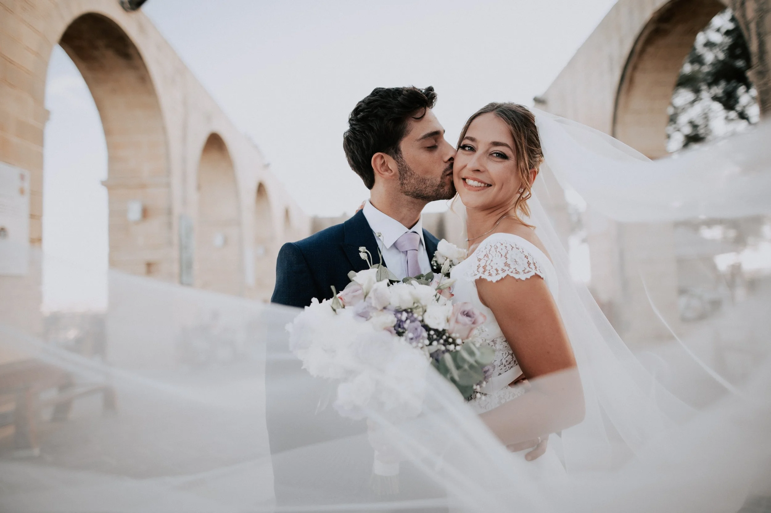 A newlywed couple, a man in a dark suit and a woman in a white lace wedding dress, sharing a kiss on their wedding day outdoors, framed by stone arches and overcast sky.