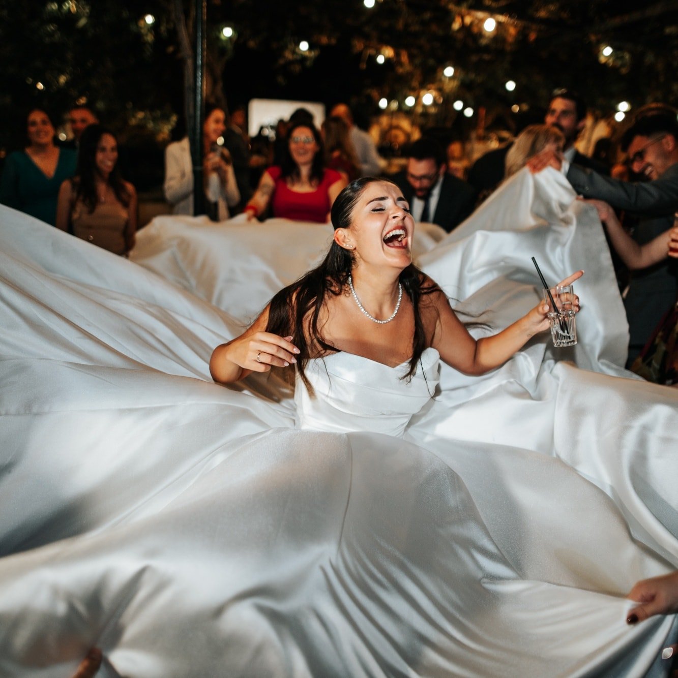 A bride in a wedding dress is laughing and holding a drink while surrounded by friends and family at her wedding reception.