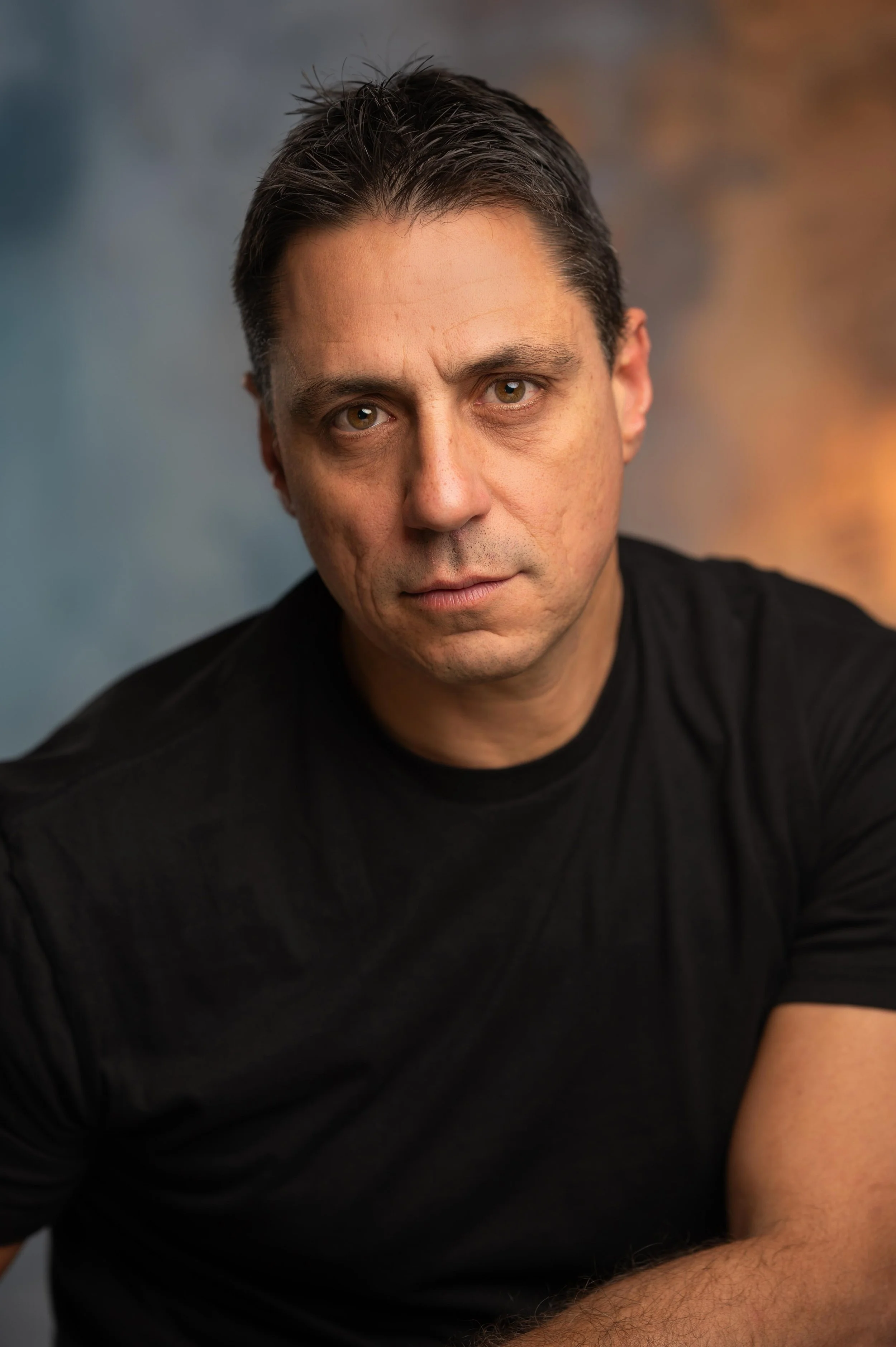 Close-up of a man with brown eyes, dark hair, and slight facial stubble, wearing a black shirt, against a blurred neutral background.