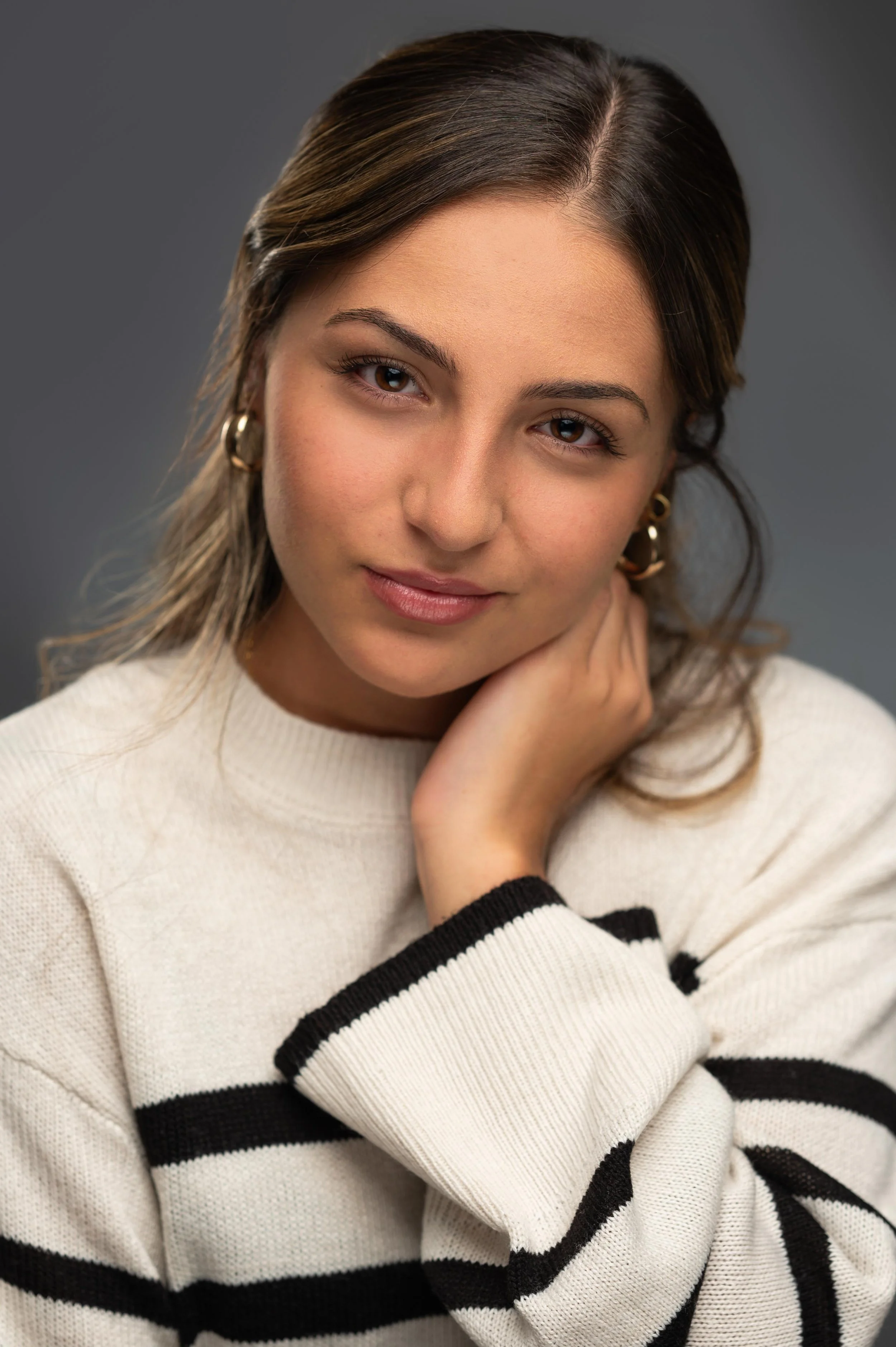 A young woman with brown hair parted to the side, wearing gold hoop earrings and a cream sweater with black horizontal stripes. She is resting her chin on her hand, looking gently at the camera with a slight smile, against a gray background.