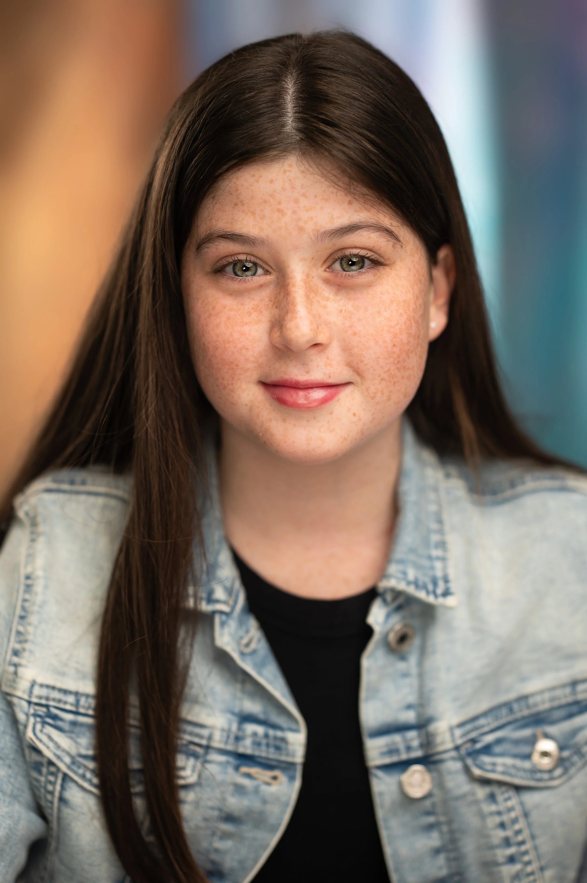 A young girl with brown hair, green eyes, and freckles, wearing a denim jacket and black shirt, smiling at the camera.