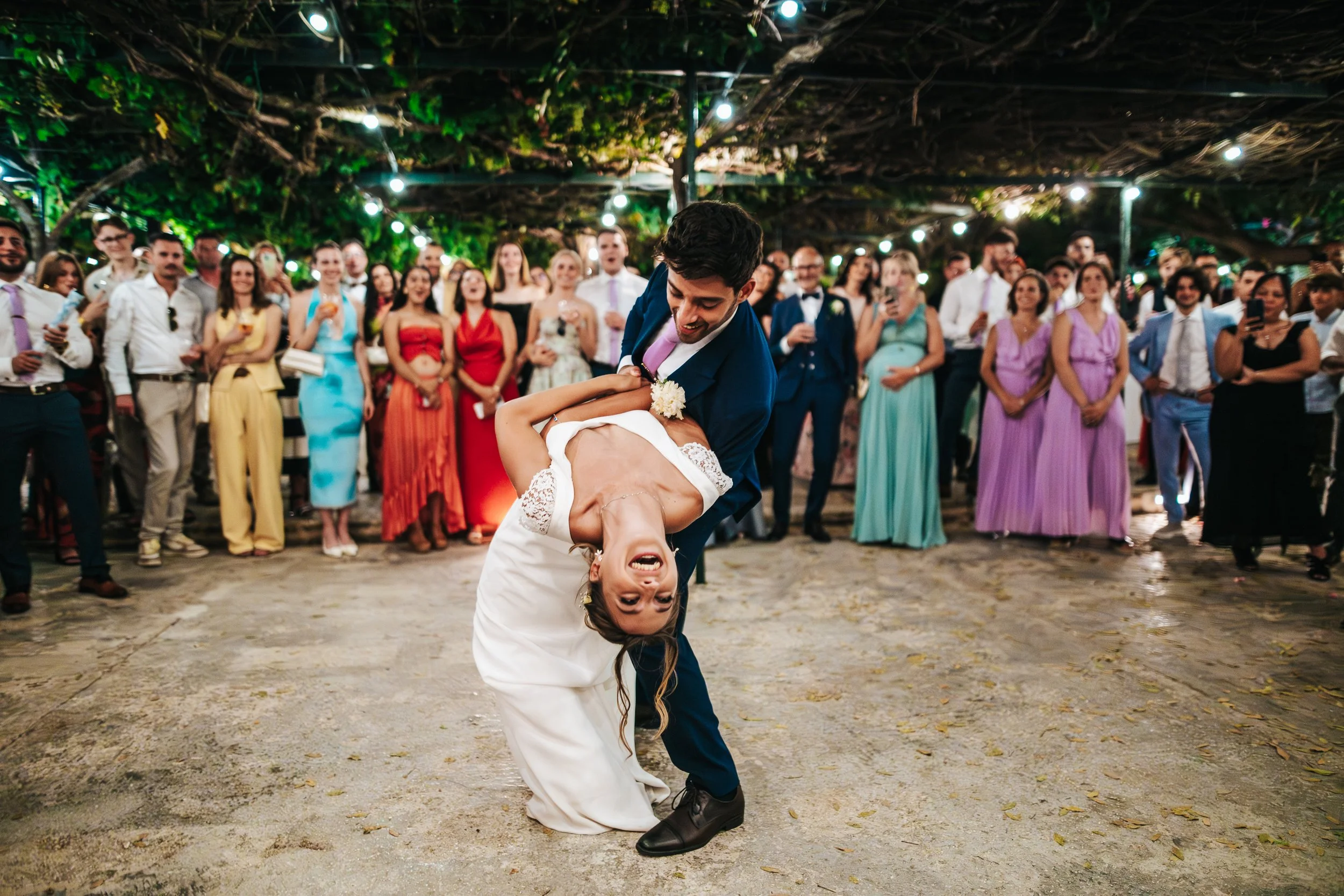 A man in a blue suit and a woman in a wedding dress are dancing at a wedding reception, with a large group of guests watching and taking pictures in a festive outdoor setting with string lights and greenery overhead.