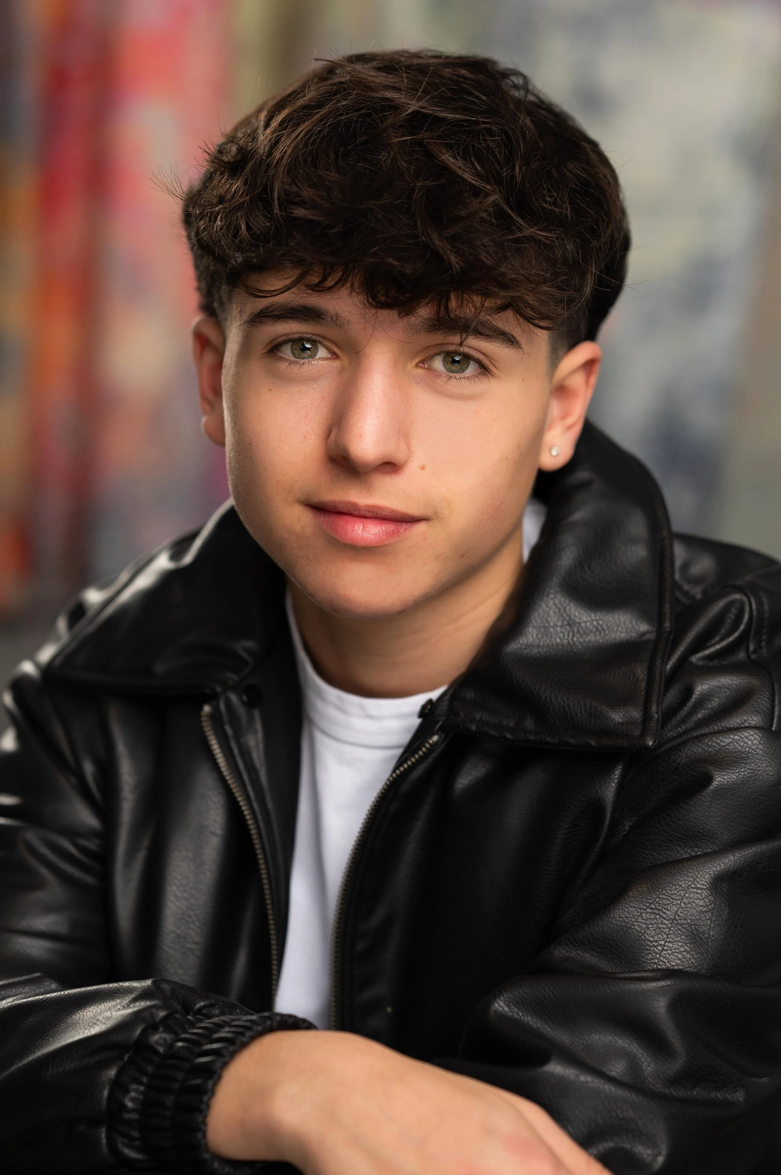 Close-up portrait of a young man with curly brown hair, green eyes, and light skin, wearing a black leather jacket and a white shirt, with a colorful, blurred background.