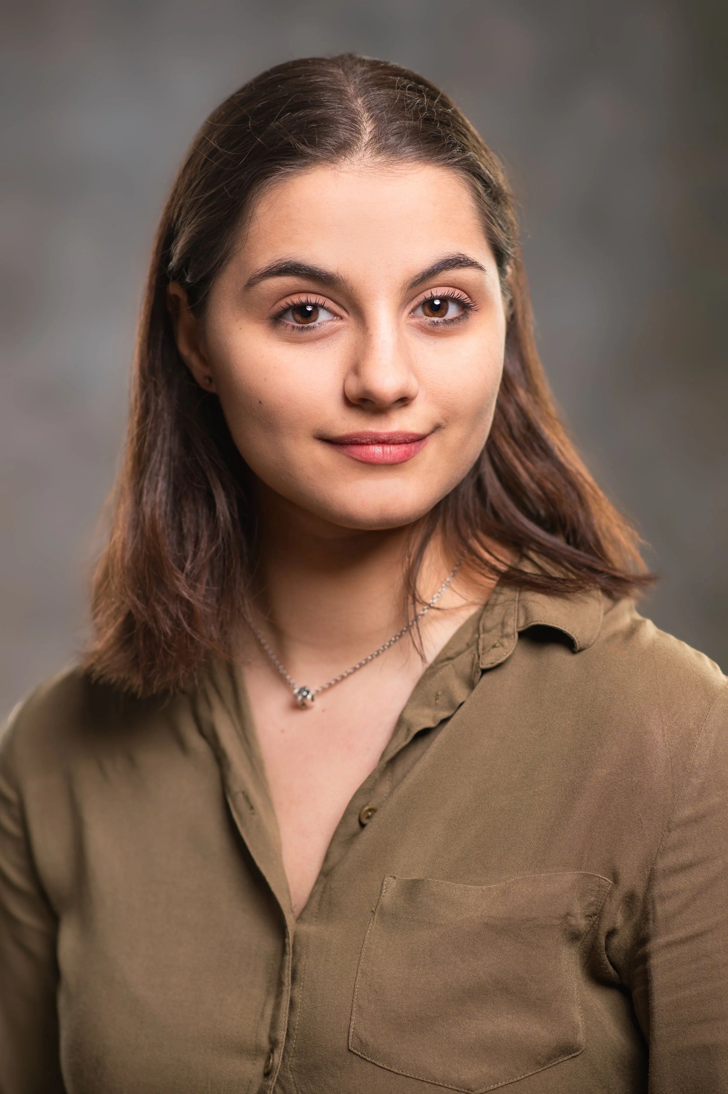 Close-up image of a young woman with brown hair, brown eyes, and light skin, smiling slightly, wearing a khaki-colored shirt and a silver necklace, with a neutral background.