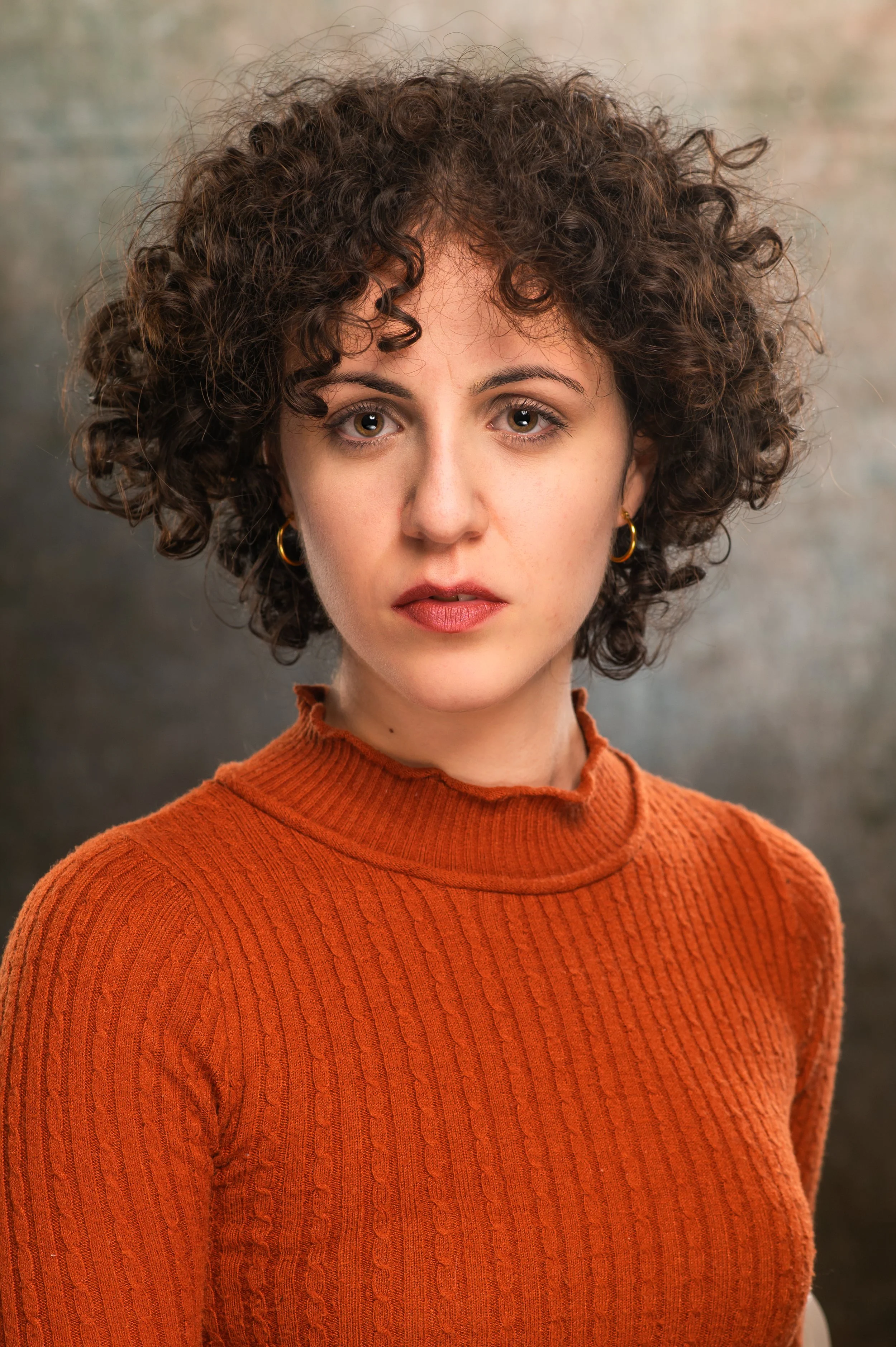 A woman with curly brown hair wearing a burnt orange ribbed turtleneck sweater, looking directly at the camera against a blurred, neutral background.