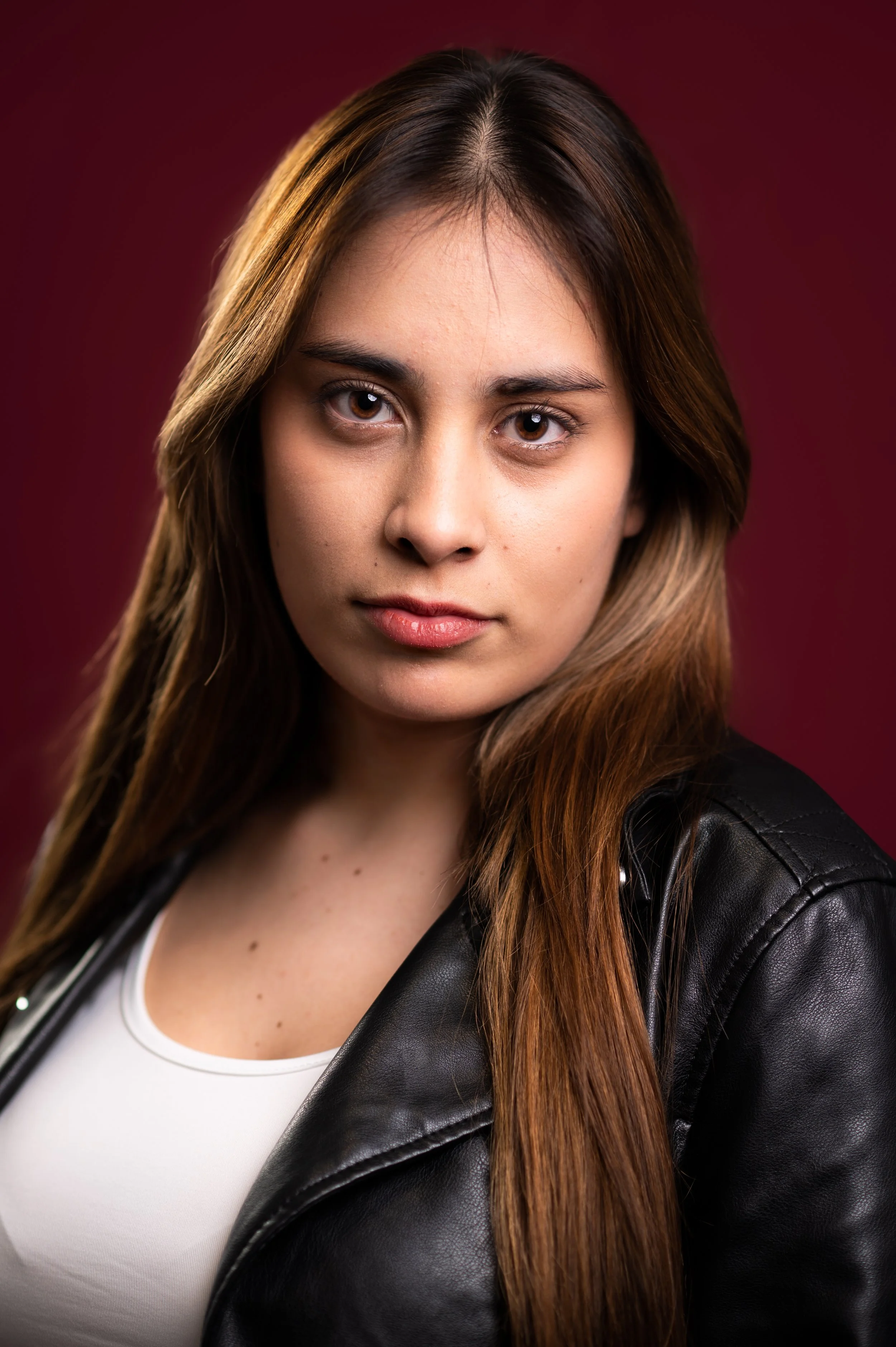 Close-up portrait of a young woman with light brown hair, wearing a white top and a black leather jacket, against a dark red background.