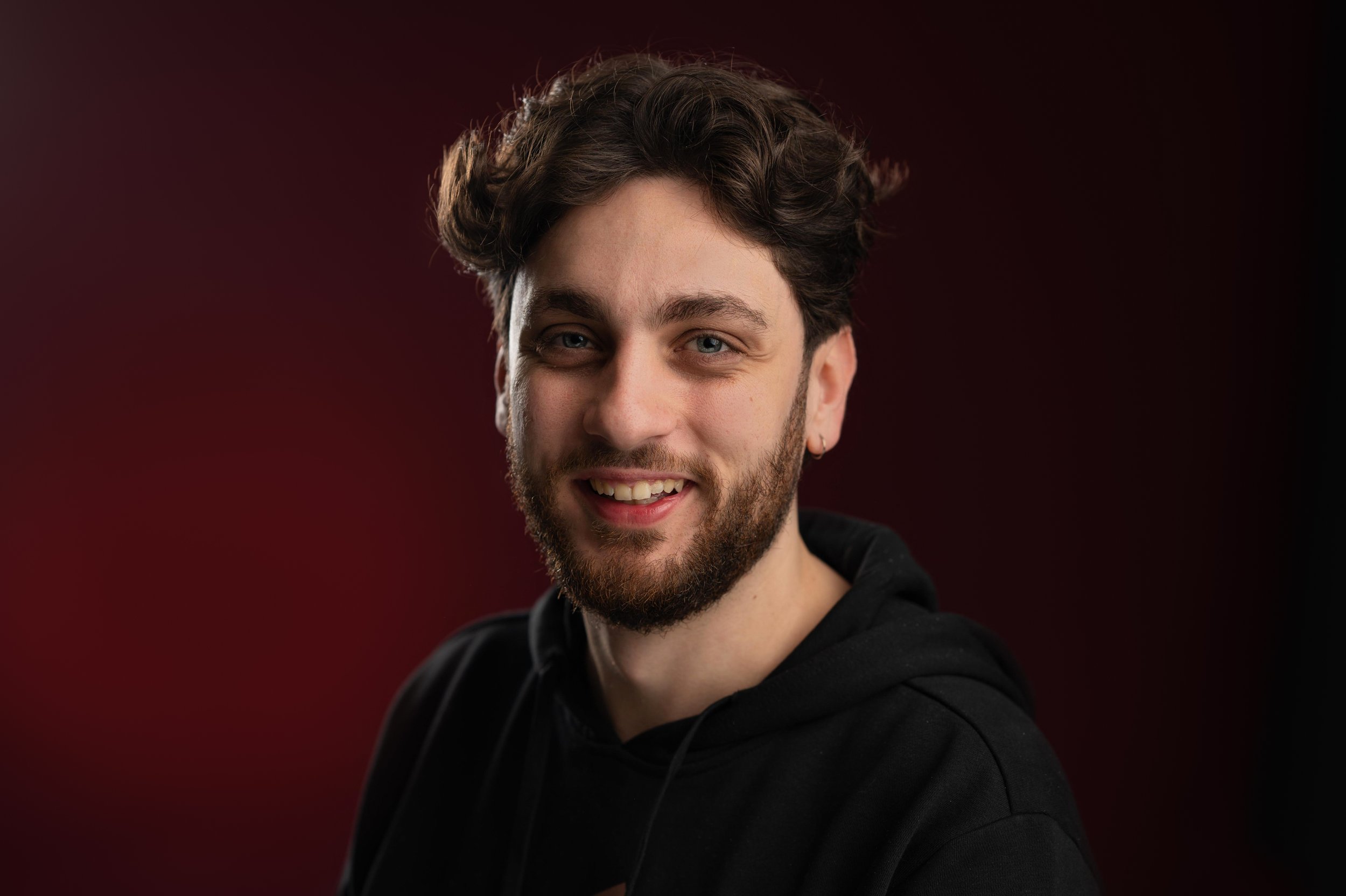 A young man with curly brown hair and a beard smiling, wearing a black hoodie against a dark red and black background.