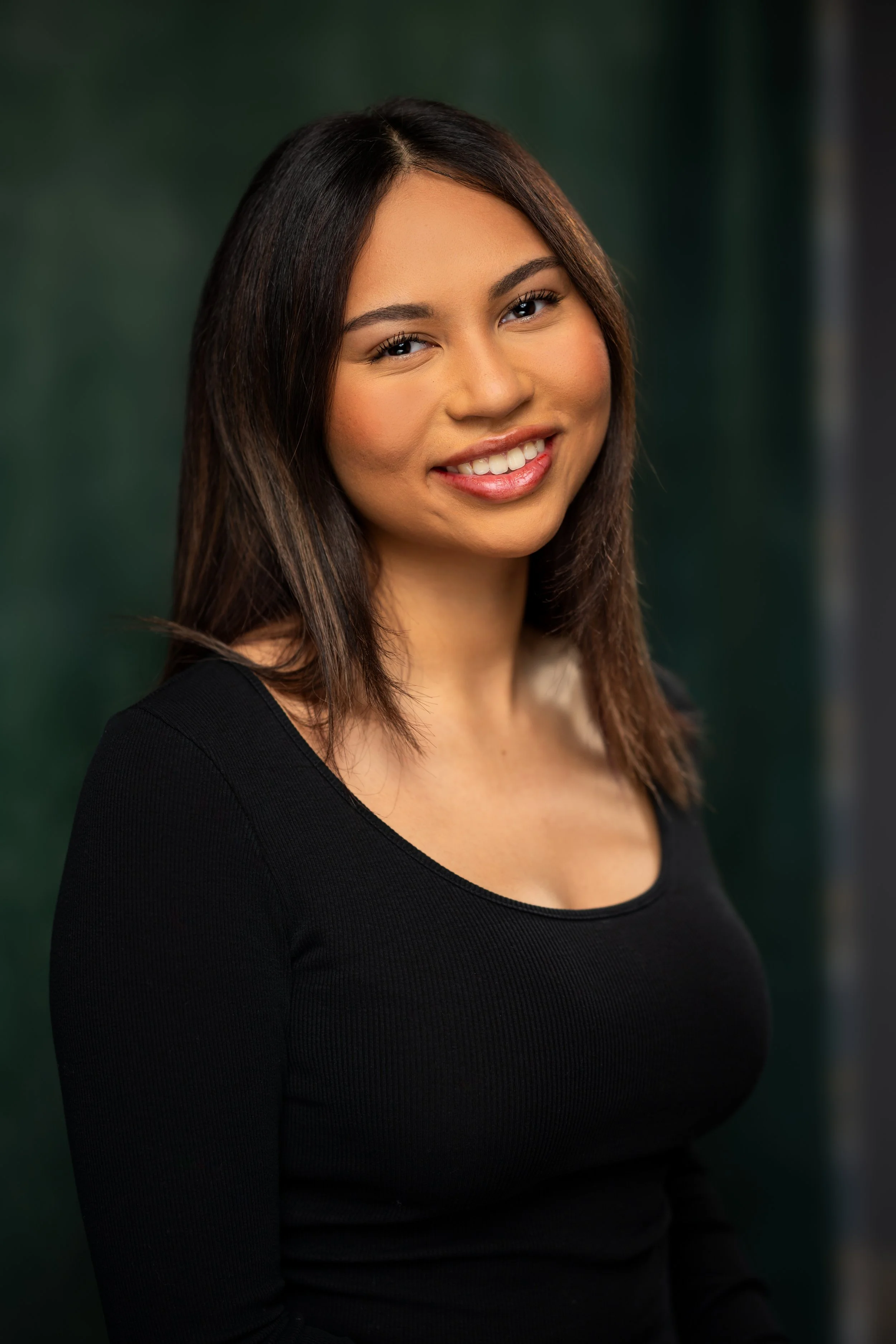 A young woman with dark brown hair and light skin, smiling and looking at the camera, wearing a black long-sleeve top, against a blurred dark green background.