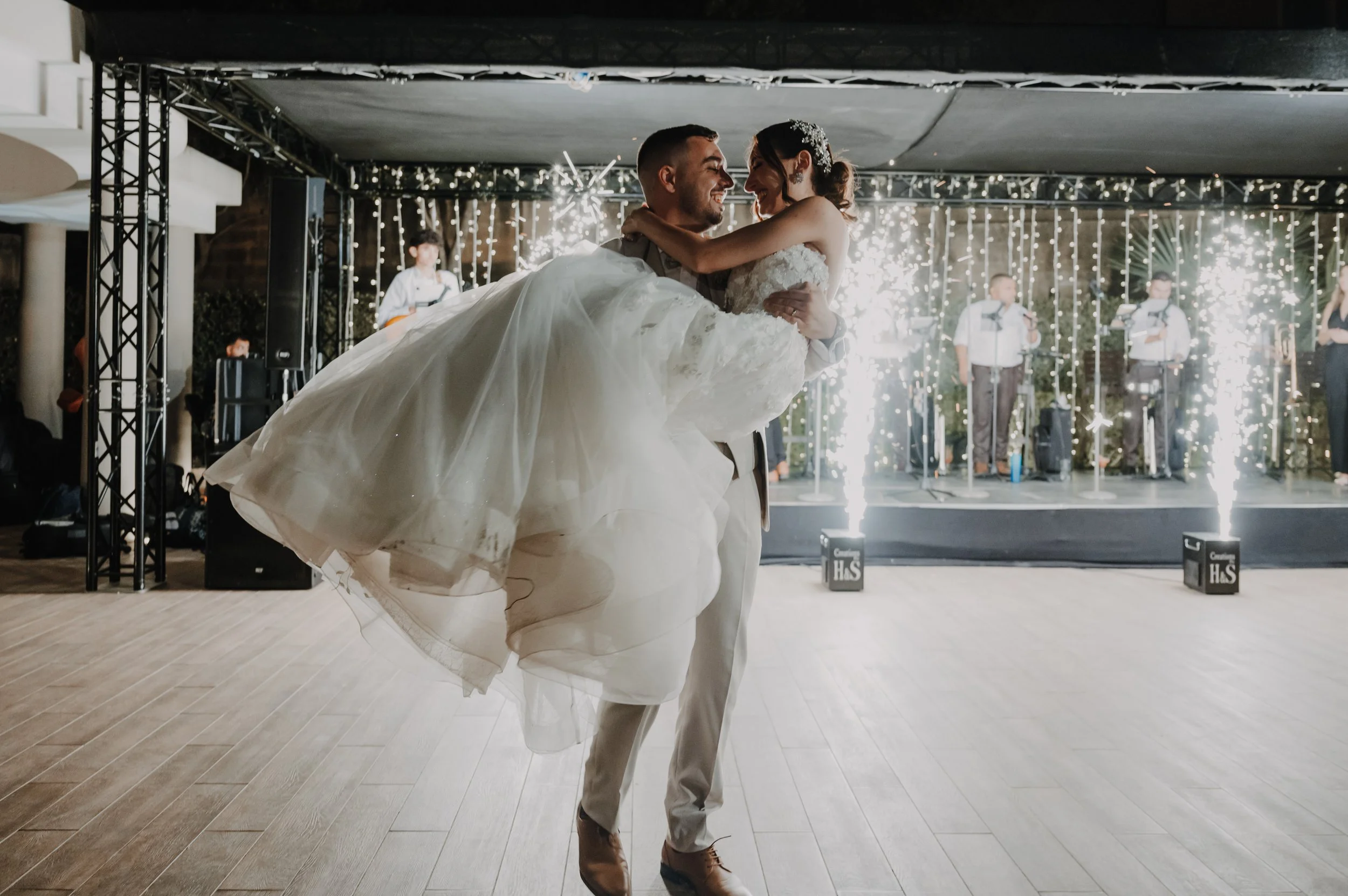 A groom holding a bride in his arms on a dance floor during a wedding reception. The bride is smiling and wearing a white wedding dress, while the groom is dressed in a beige suit. Sparkler fountains are visible in the background, along with musician