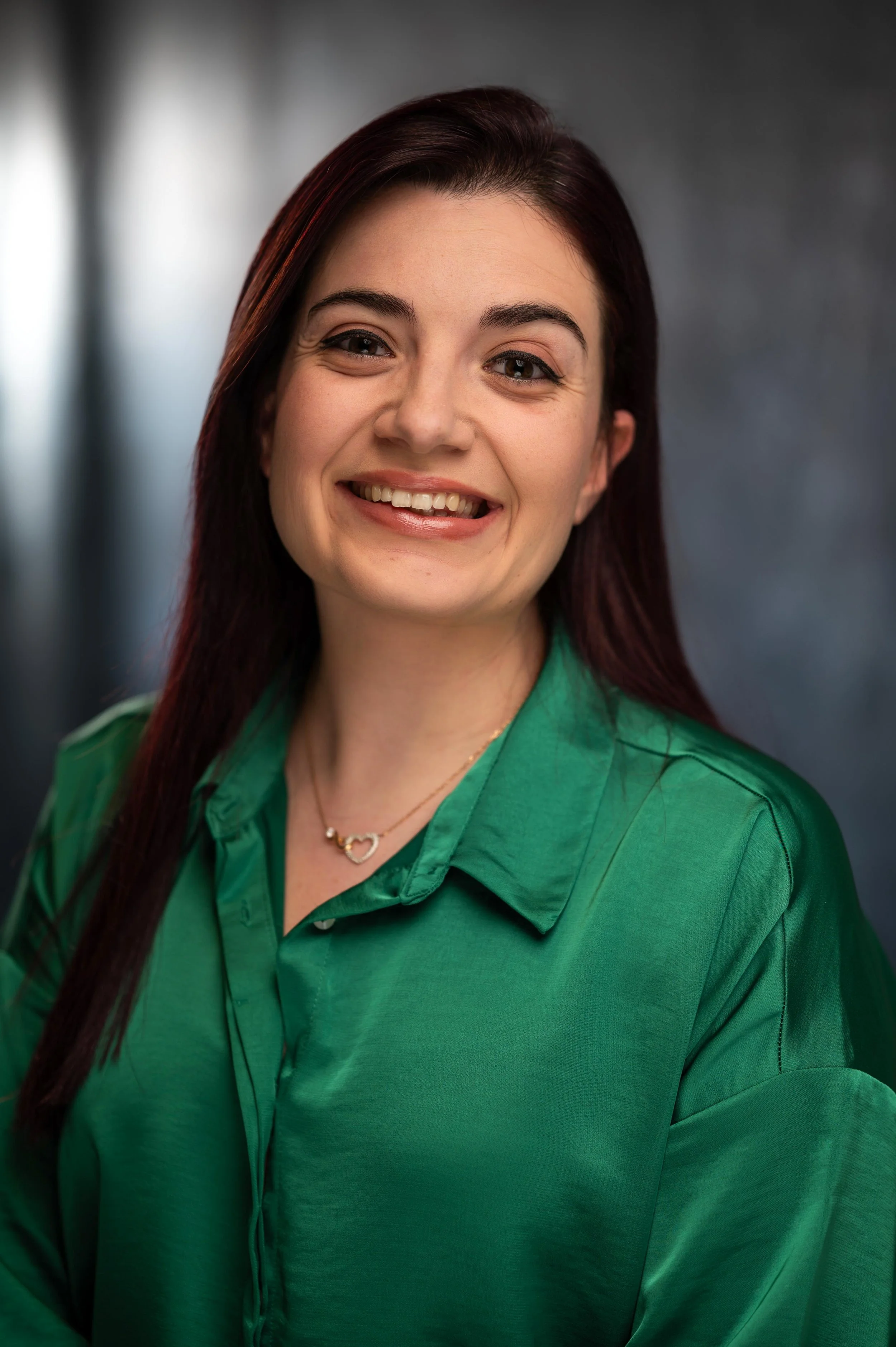 A smiling woman with dark red hair, wearing a green button-up shirt and a heart-shaped necklace, standing in front of a blurred gray background.