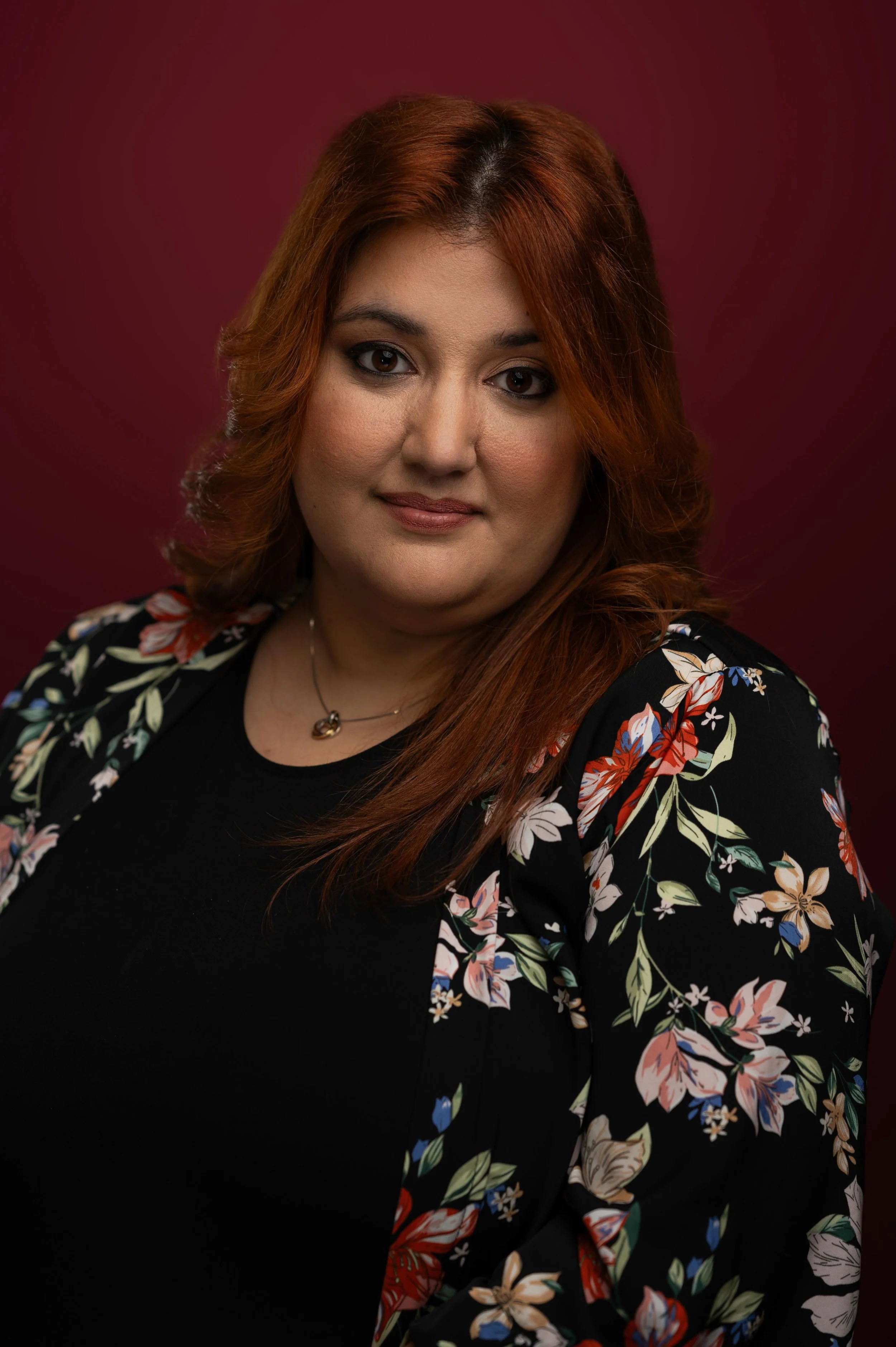 A headshot of a woman with long, wavy red hair, wearing a black top and a floral jacket with colorful flowers, against a dark red background.