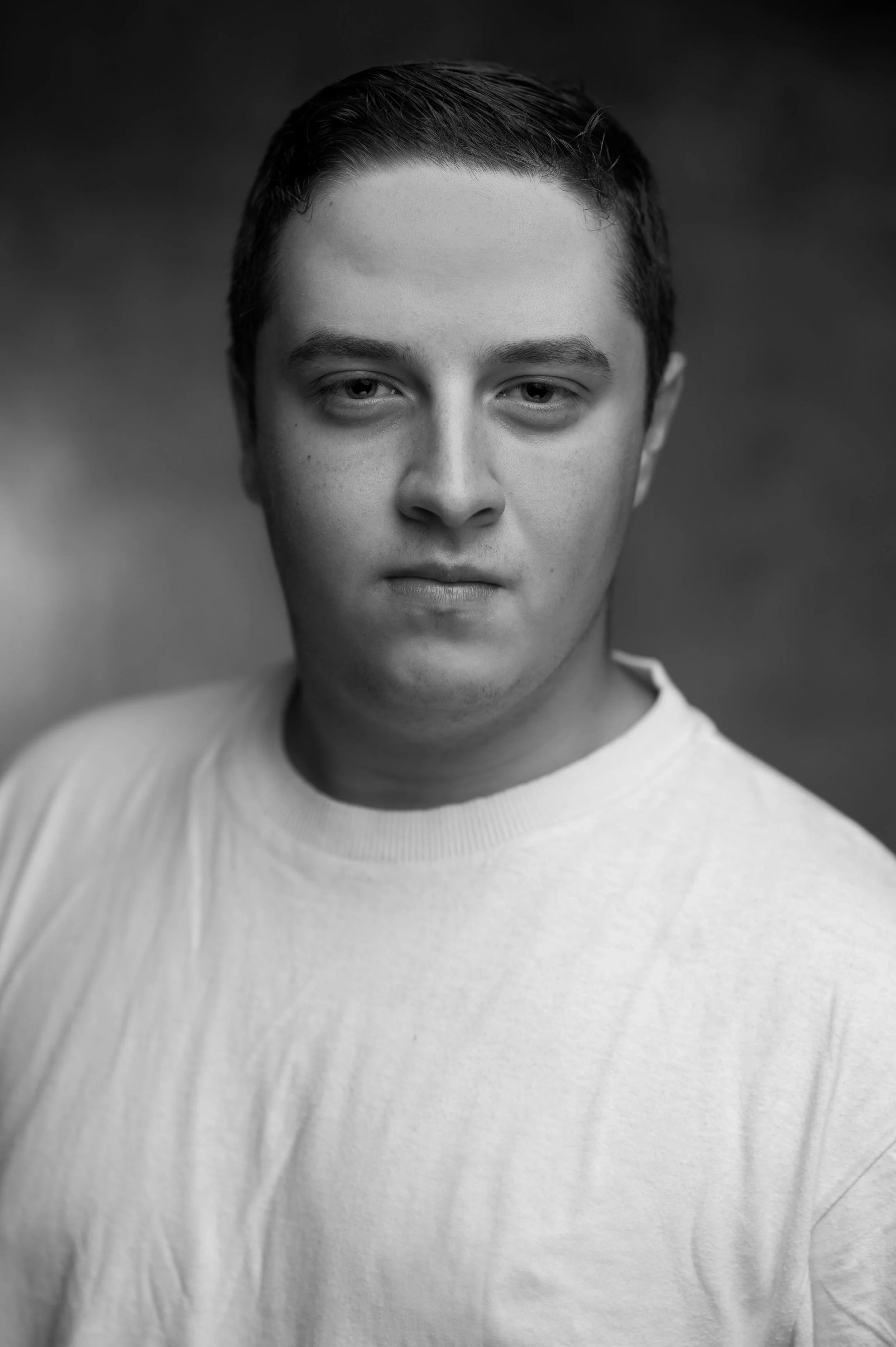Black and white portrait of a young man with short, dark hair, wearing a light-colored shirt, looking directly at the camera with a neutral expression.