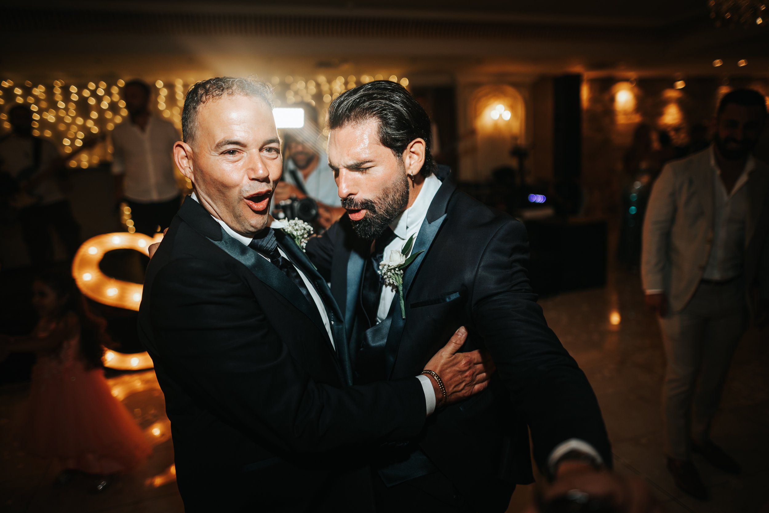 Two men in tuxedos warmly embrace and dance at a wedding reception, with warm golden lighting and blurred background of guests and decorations.