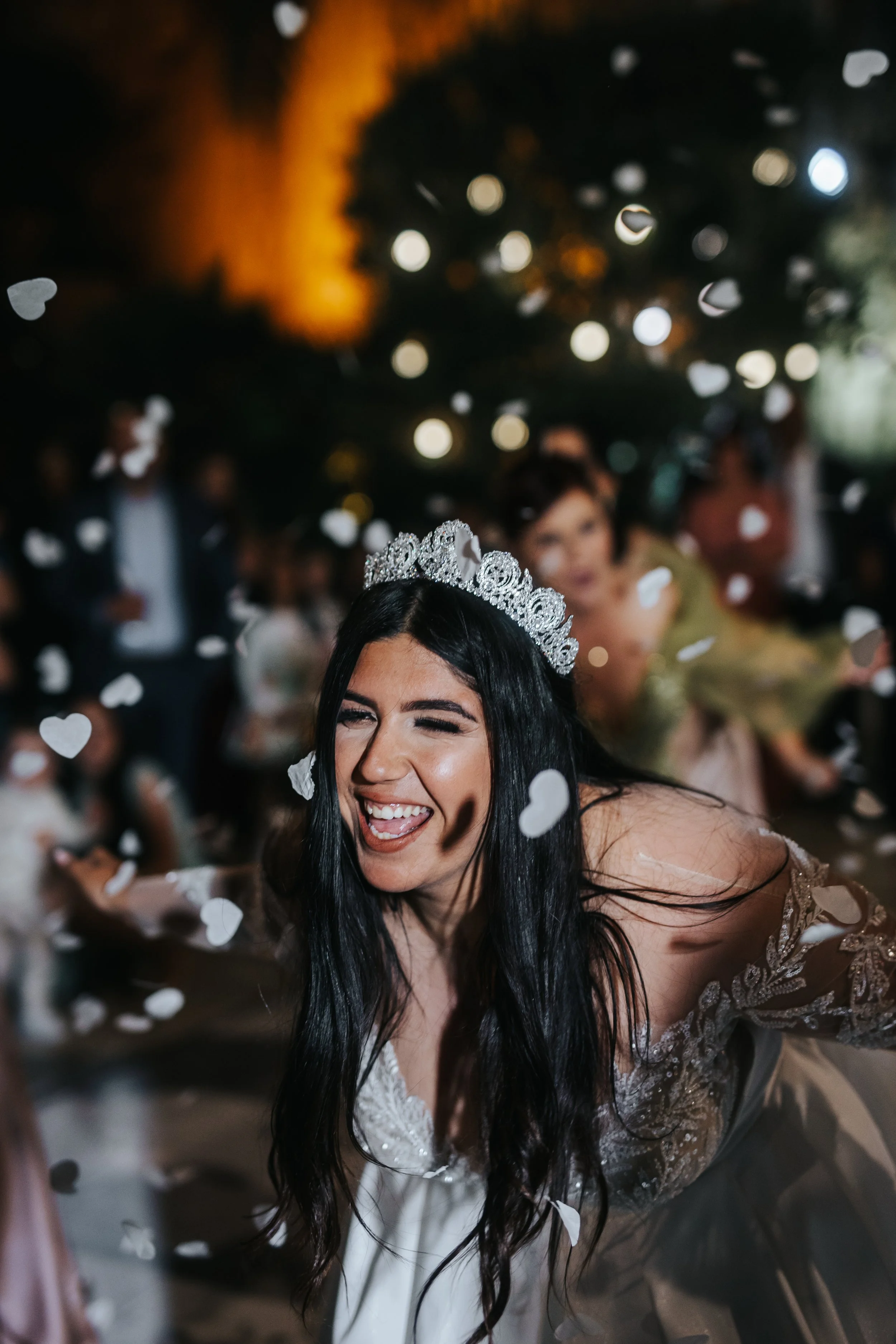 A woman wearing a tiara smiles joyfully while throwing white confetti at a celebration event. There are blurred people and lights in the background.