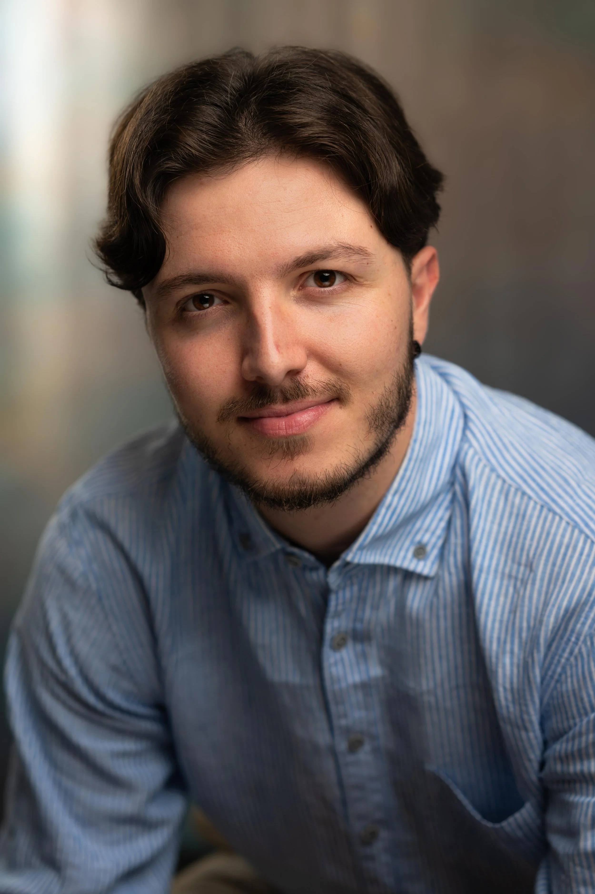 A young man with dark hair, trimmed beard, and wearing a blue striped shirt, looking directly at the camera with a slight smile.